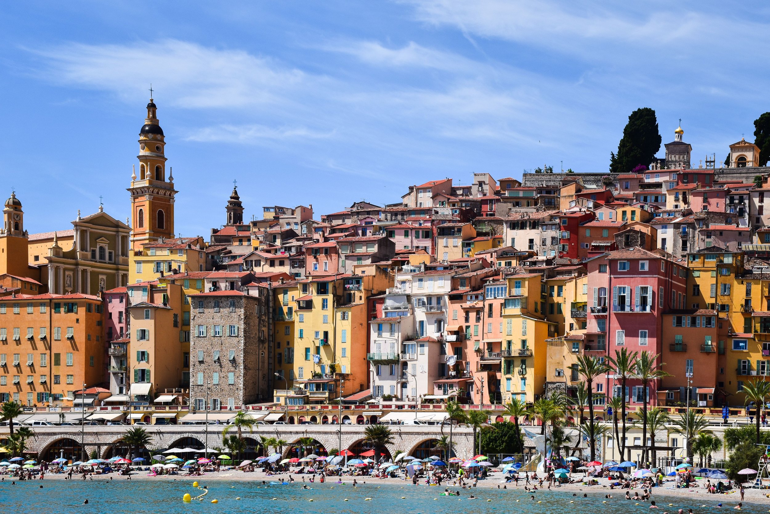 Colorful hillside buildings with church steeples, overlooking a beach with umbrellas and people, under a blue sky with clouds.