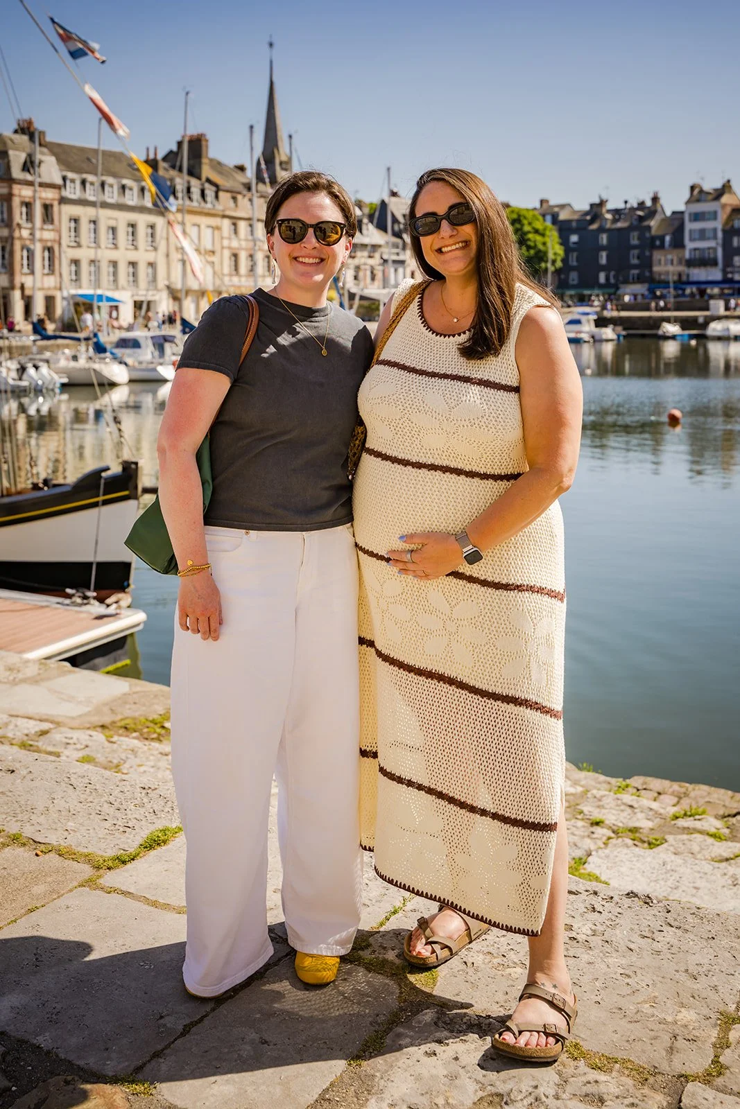 Two women smile by a sunny harbor while vacationing in France with travel company Maison d'Amies.