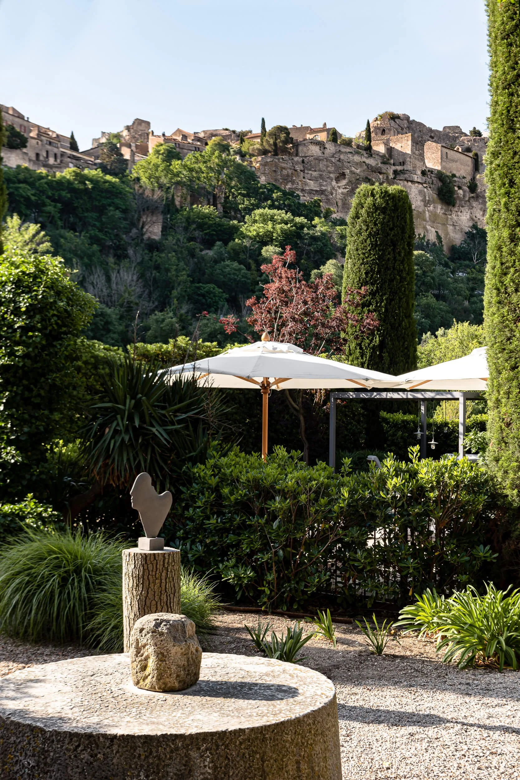 A garden scene with lush greenery, a tree with pink blossoms, and a white patio umbrella. There is a stone sculpture of a chicken on a stump, surrounded by rocks and various plants. In the background, tall cypress trees and a hillside with historic stone buildings and ruins are visible.