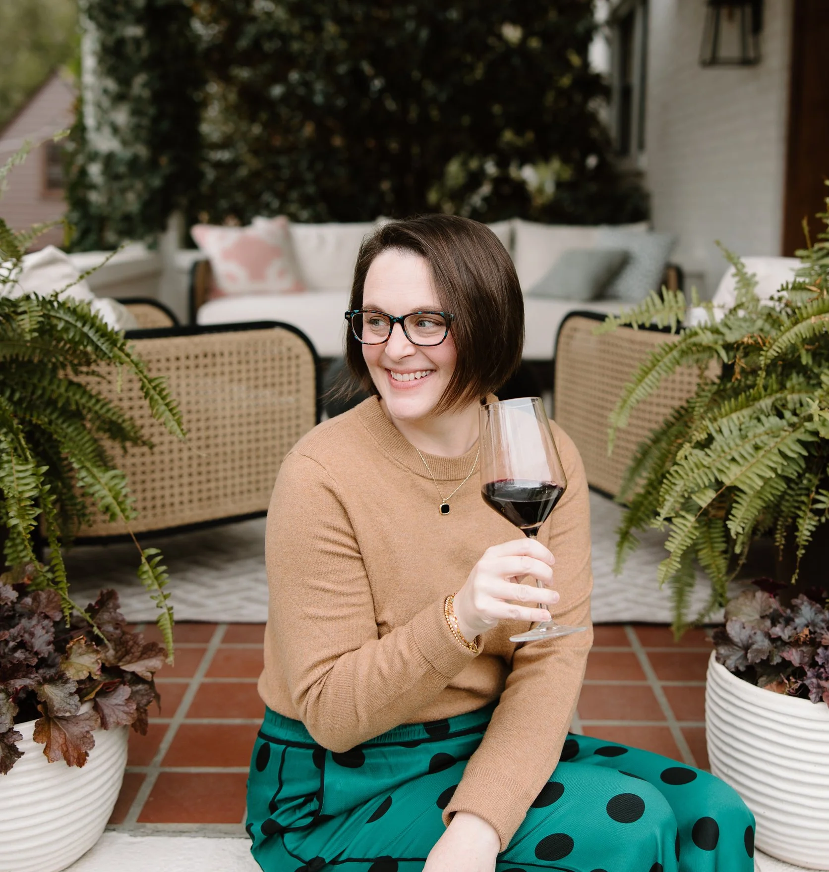 A woman with short brown hair, glasses, and a tan sweater sitting outdoors, holding a glass of red wine, smiling, with potted plants and outdoor furniture in the background.