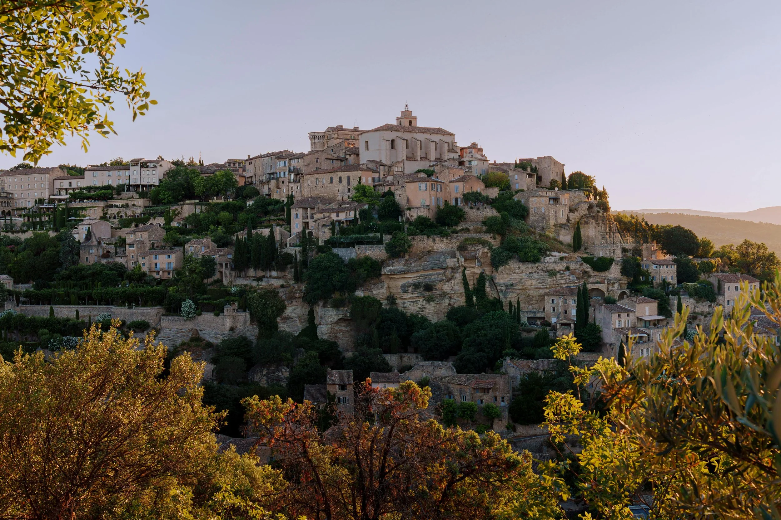A hillside village with historic stone buildings and a large church at the top, surrounded by lush green trees, with the sunlight casting a warm glow on the scene.
