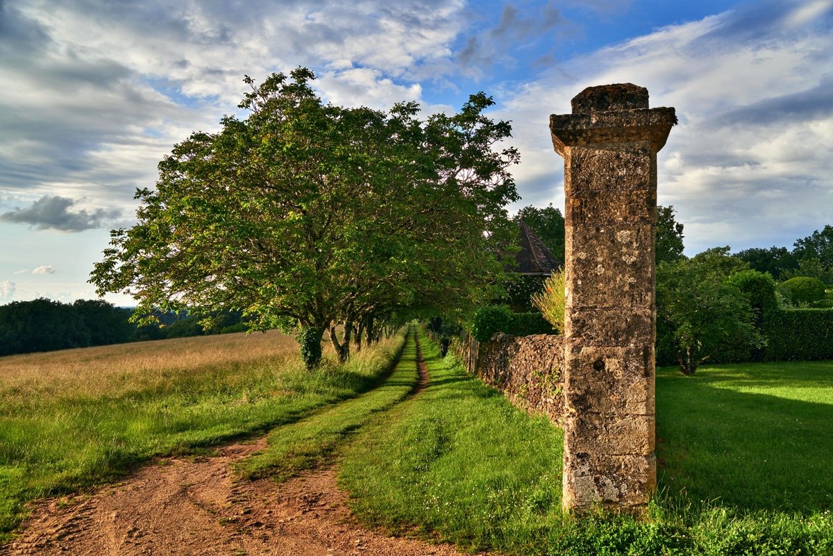 A dirt pathway runs alongside a stone wall with tall trees providing shade, in a lush green landscape under a partly cloudy sky.