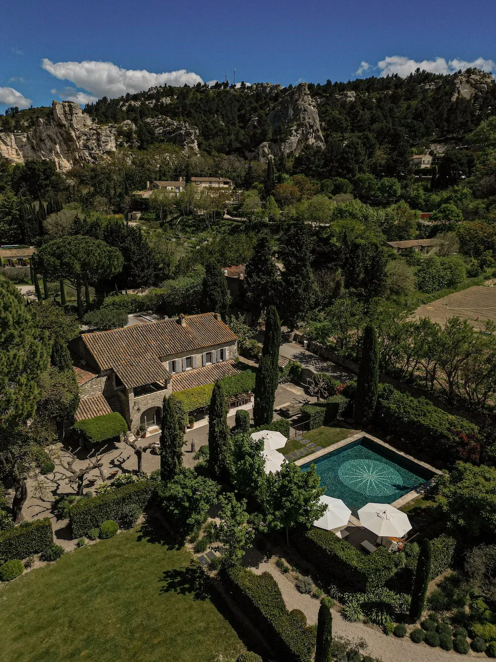 A hillside estate with Mediterranean-style house, swimming pool, and patio area, surrounded by lush trees and mountains under a blue sky.