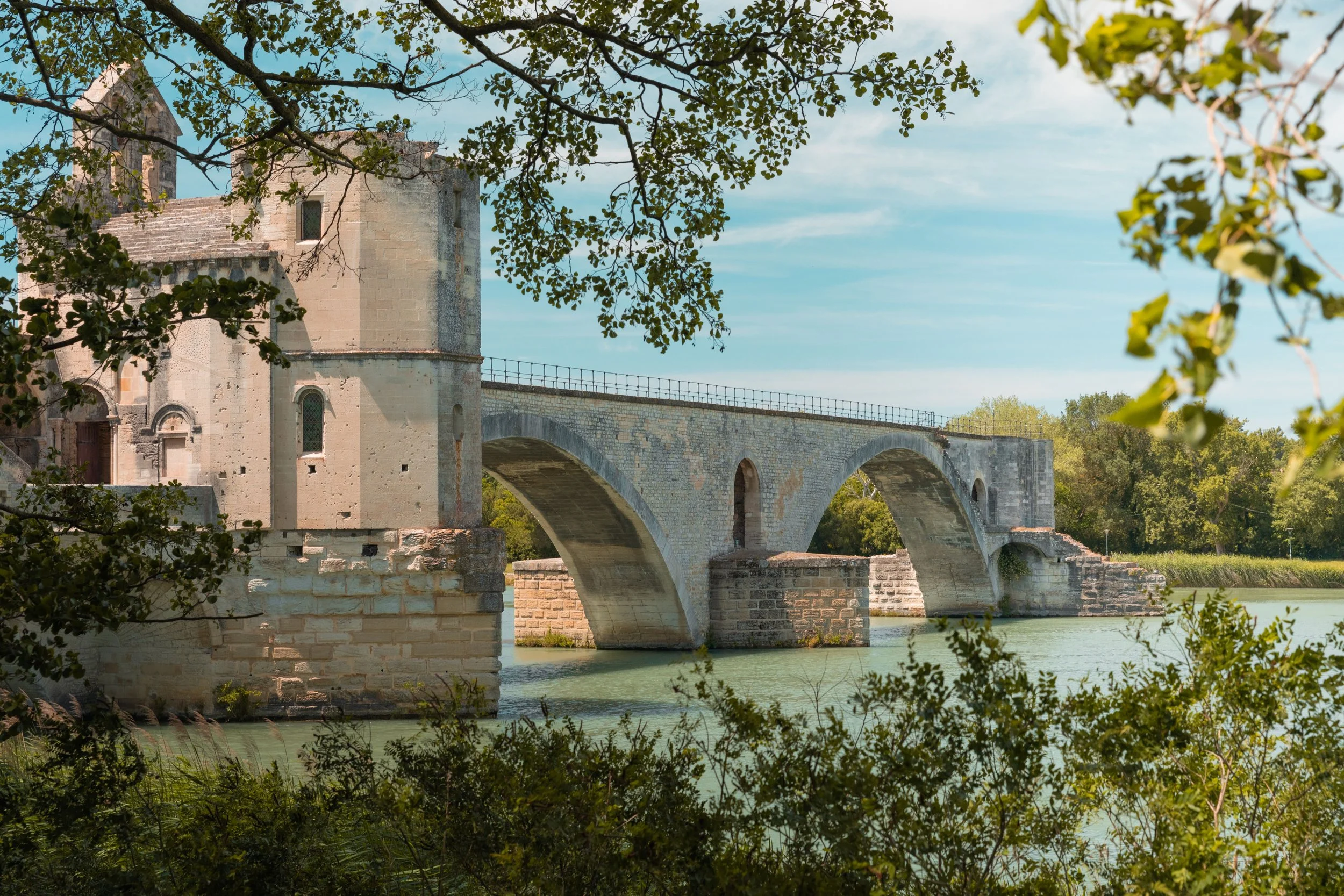 Stone bridge over a river with a historic stone building and trees in the background, seen through foliage.