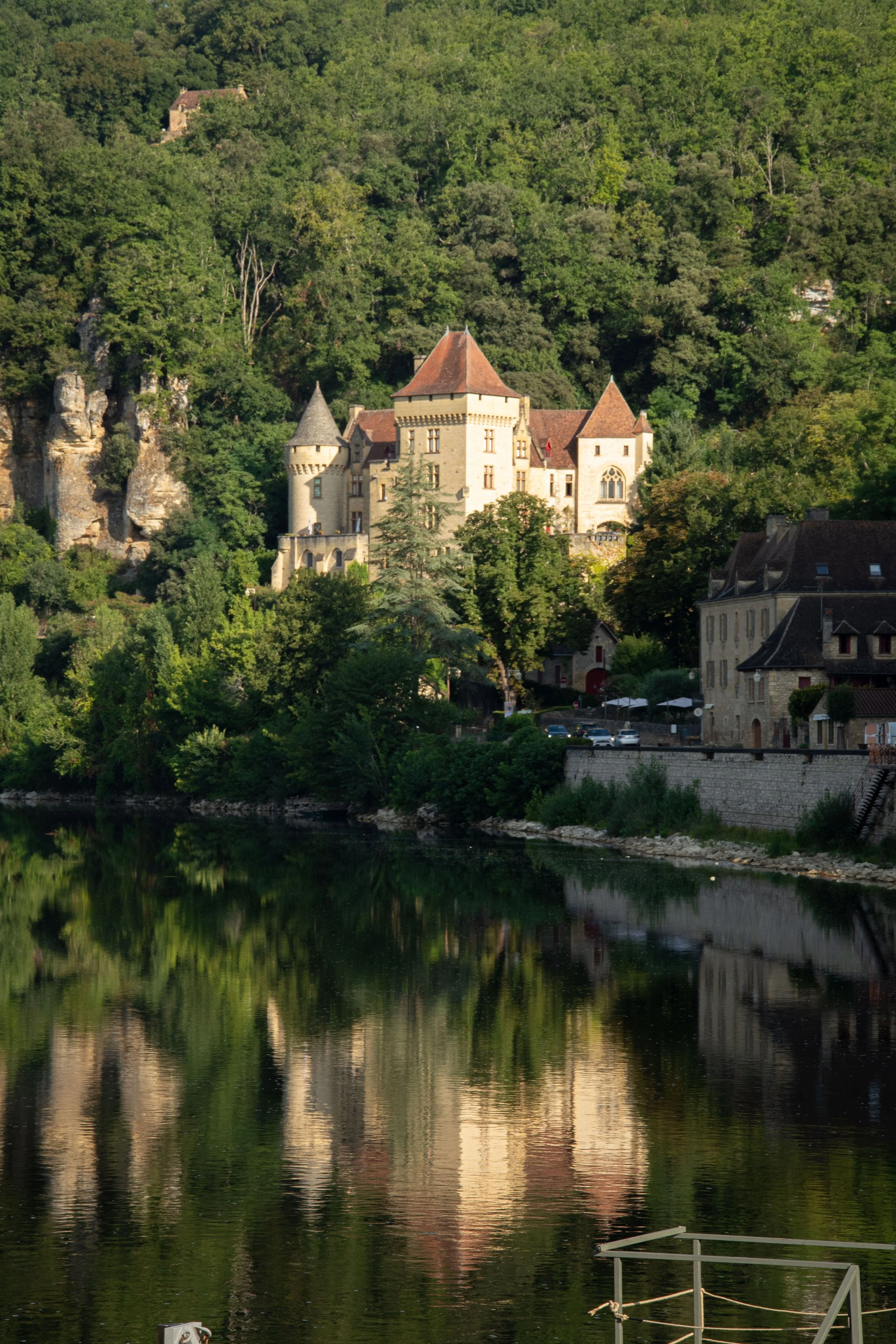 A castle with towers and a red-tiled roof surrounded by dense green trees situated at the edge of a river, with reflections of the castle and trees visible in the water.