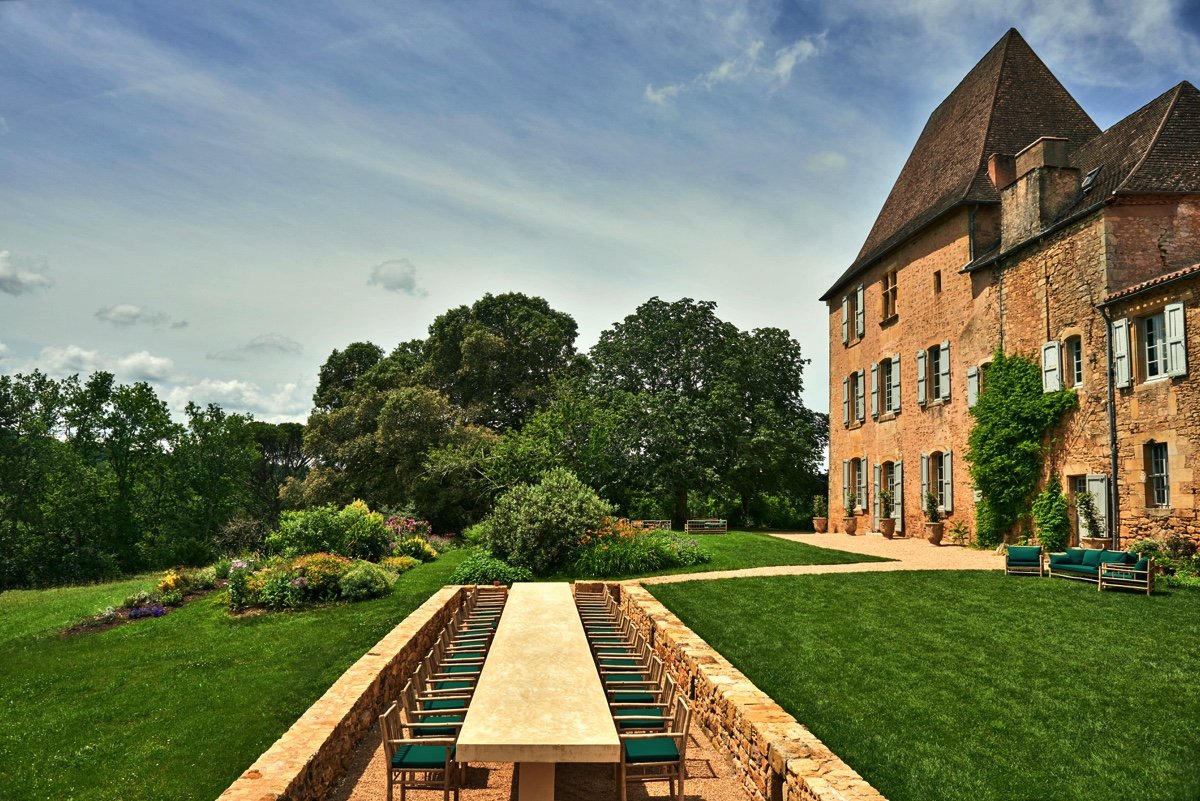 An outdoor scene featuring a long rectangular table with chairs on a brick patio, adjacent to a large old stone house with multiple windows, surrounded by a well-maintained lawn, trees, and flowering shrubs under a partly cloudy sky.