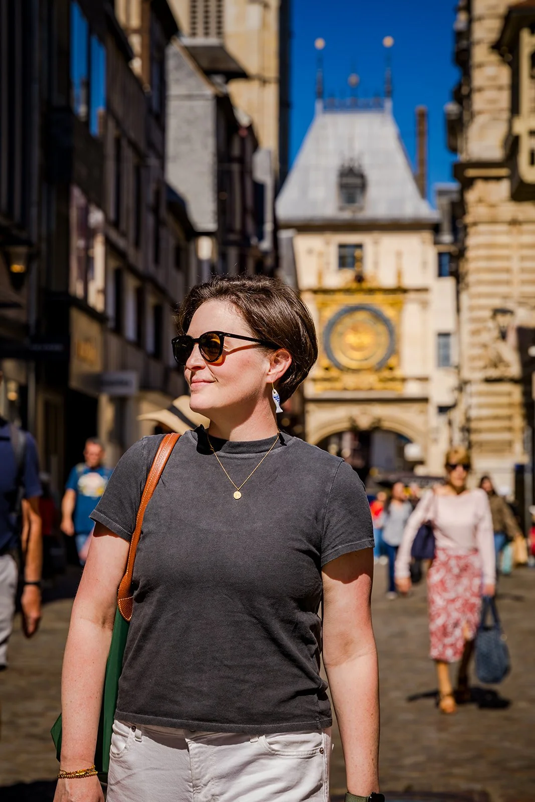 A woman with short dark hair and sunglasses standing in a busy city square with historic buildings and clock tower in the background.