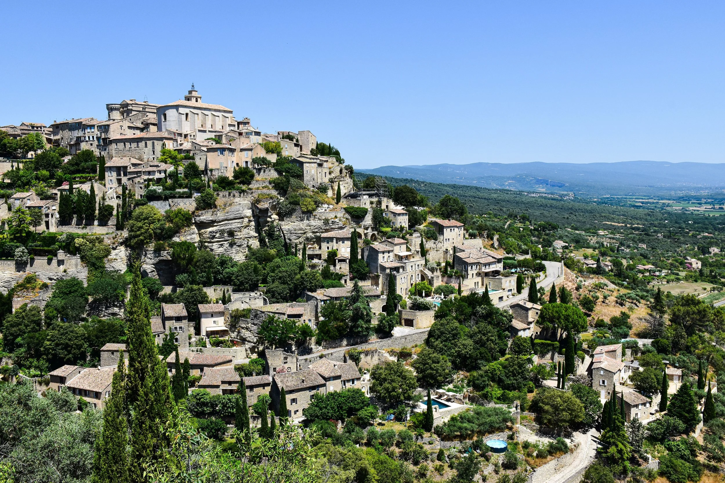 A hillside village with historic stone buildings and lush greenery, overlooking a vast plain with mountains in the distance under a clear blue sky.