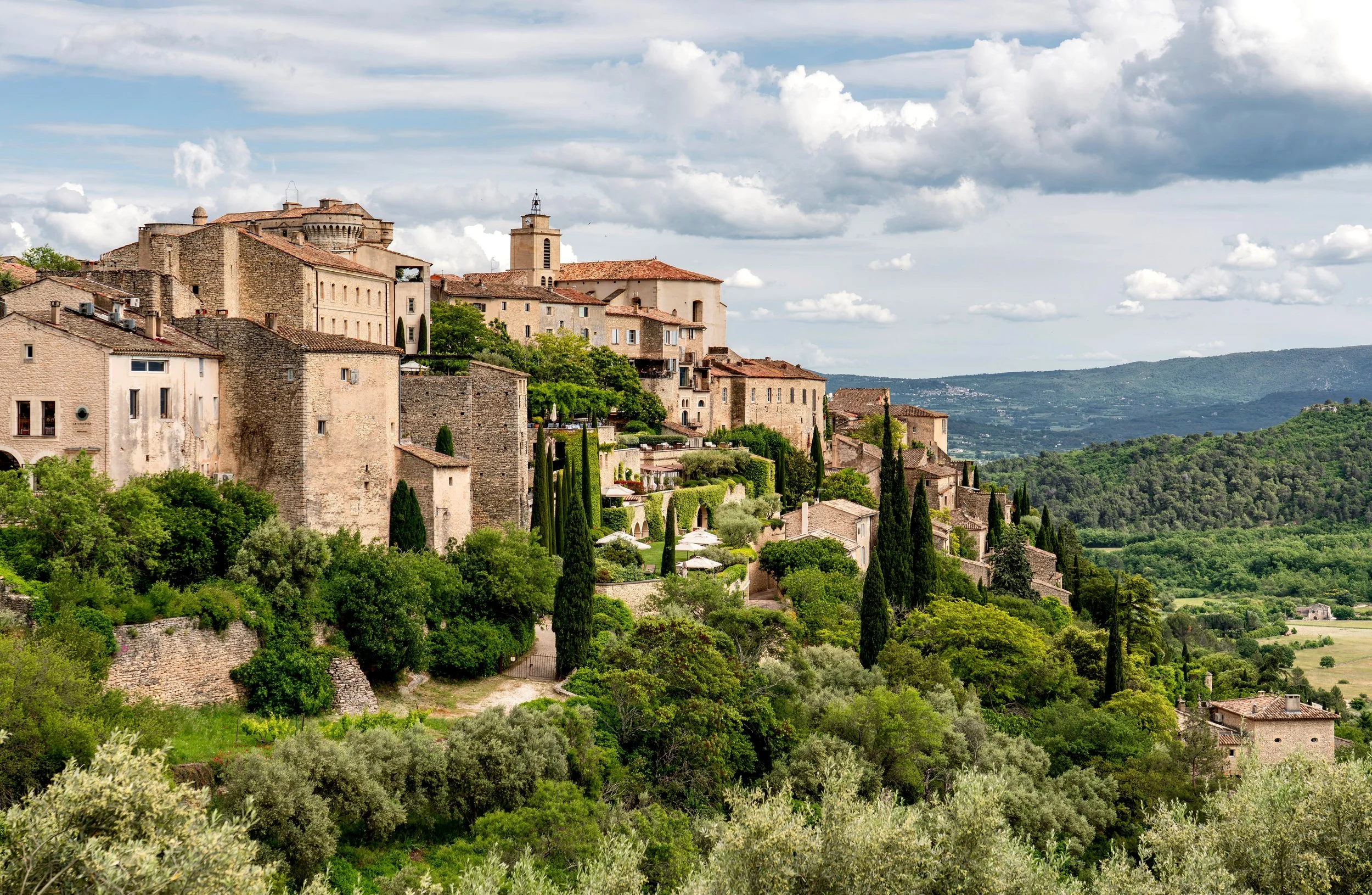 A hillside view of a historic European town with stone buildings, red-tiled roofs, and a church steeple, surrounded by lush greenery and distant hills under a partly cloudy sky.