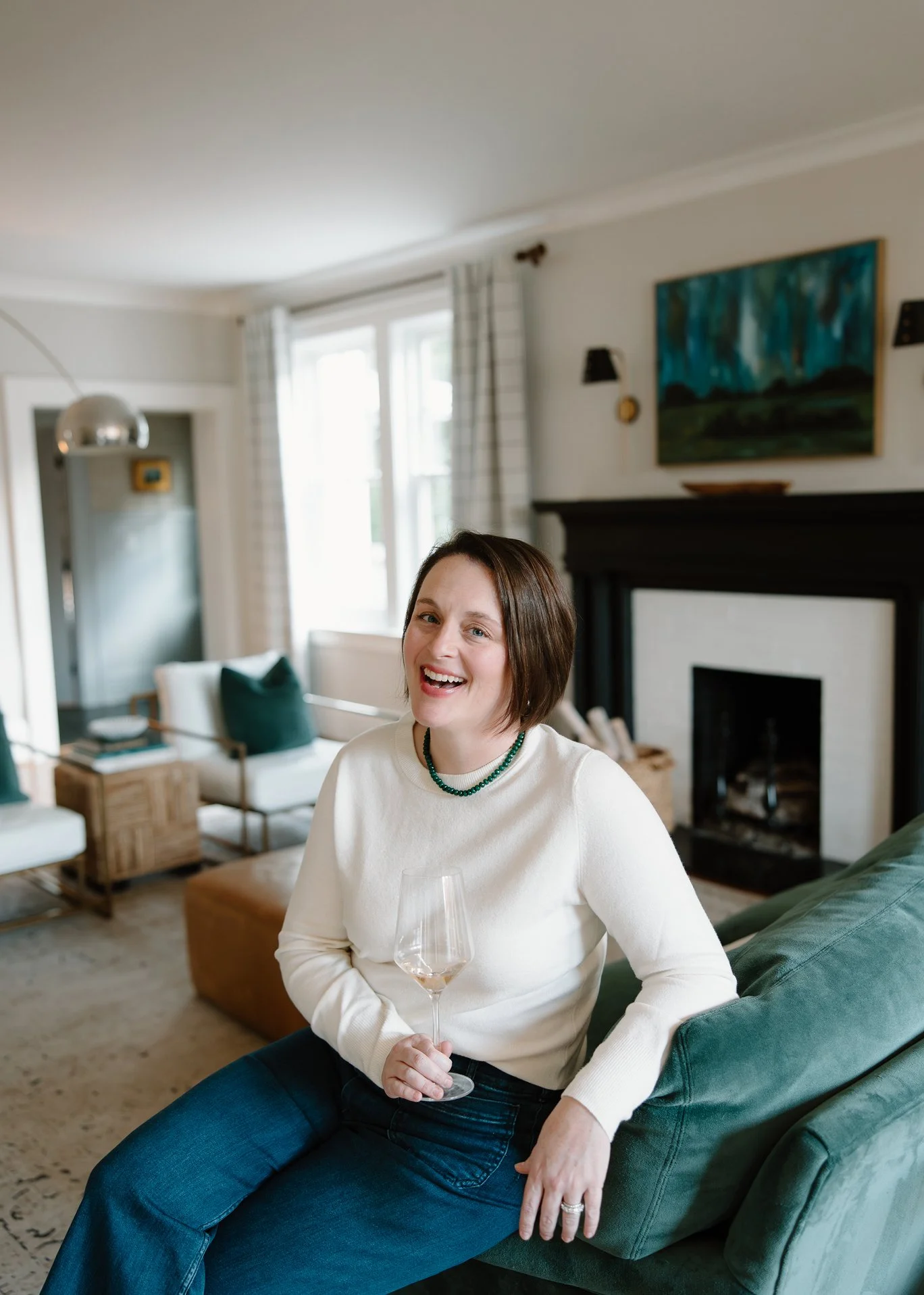 Woman sitting on a green couch holding a glass of white wine, smiling in a well-lit living room with artwork, fireplace, and modern furniture.