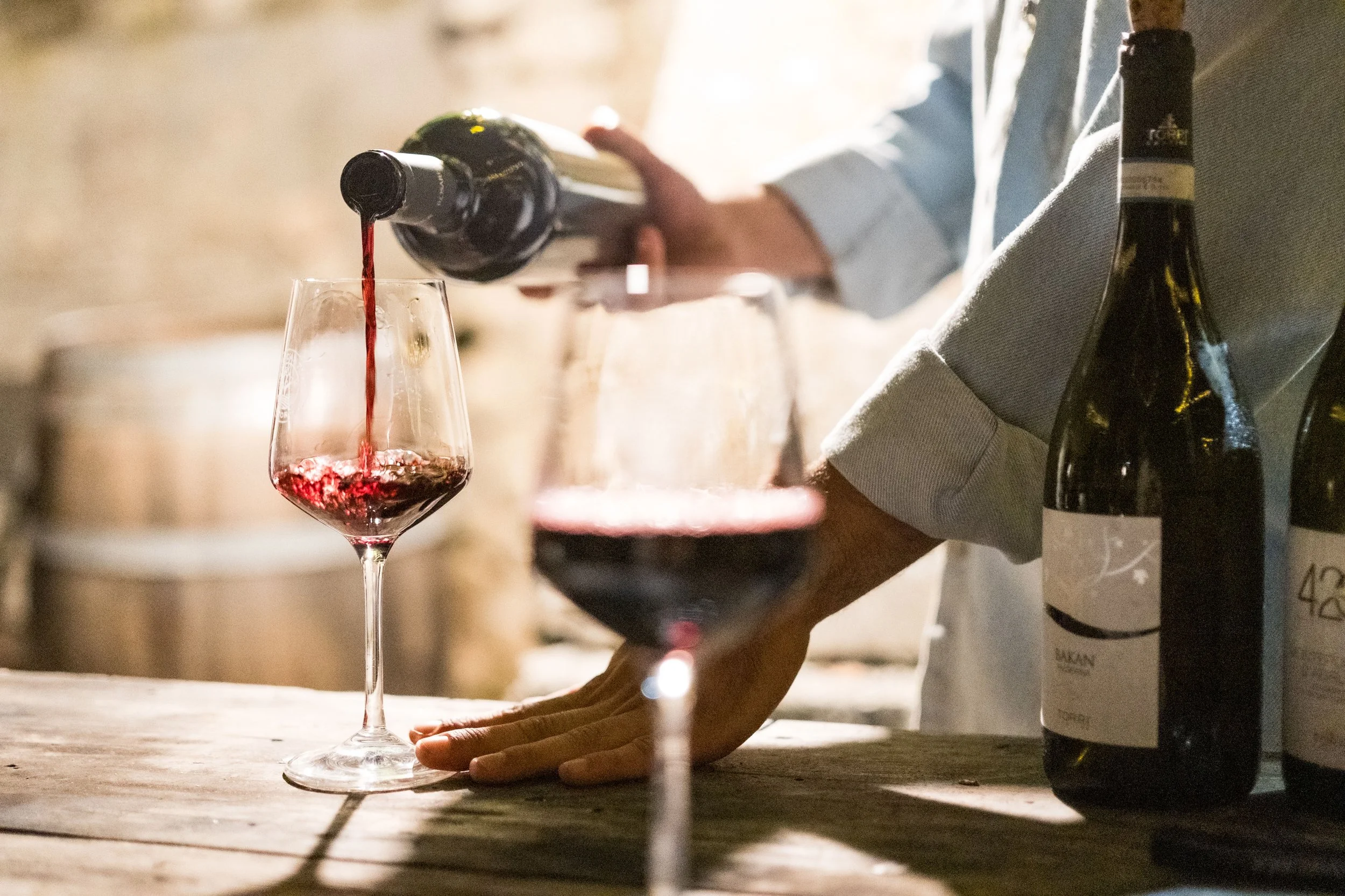 A person pouring red wine into a wine glass on a wooden surface, with wine bottles nearby. The scene appears to be in a rustic or cellar setting.