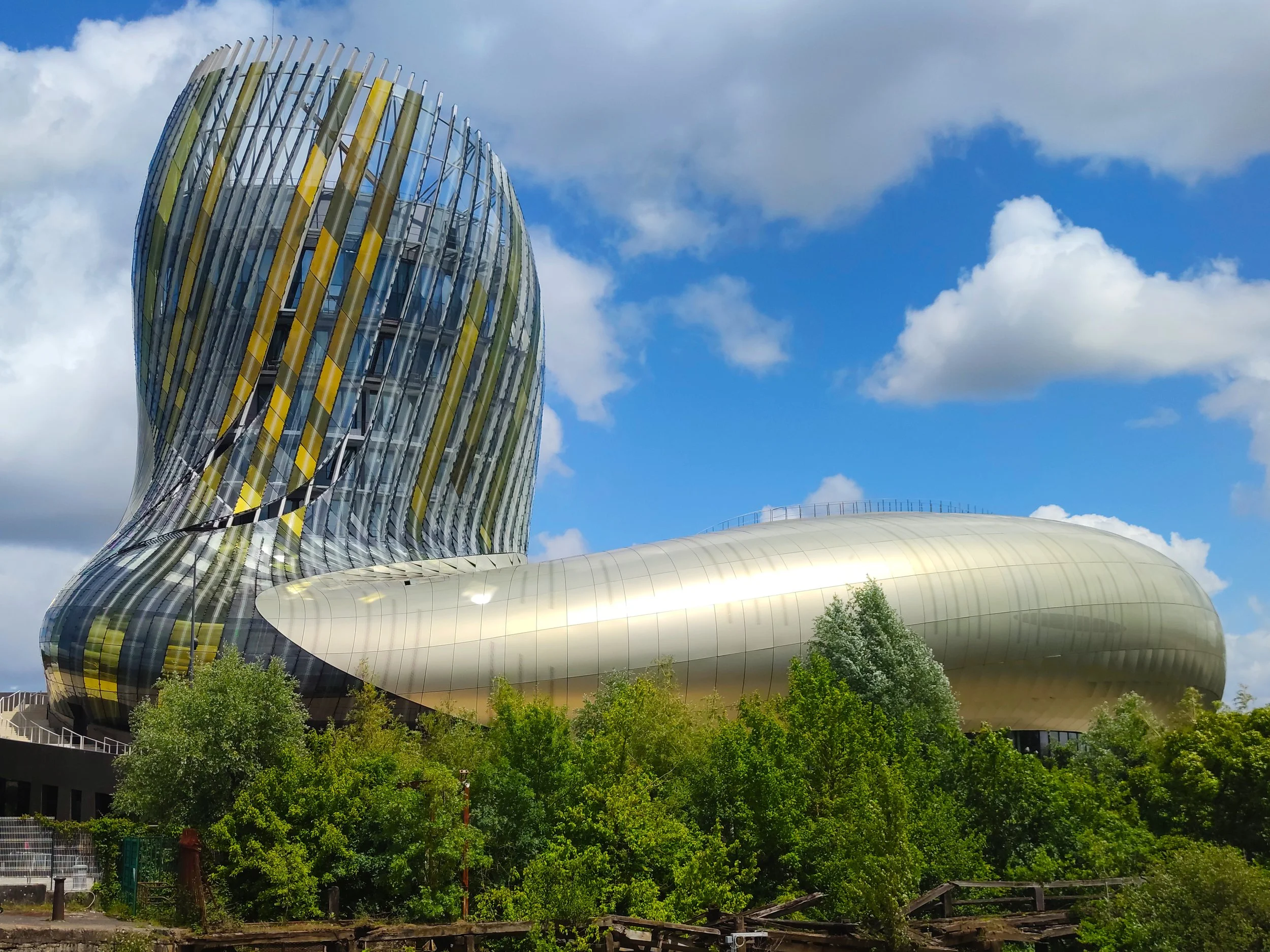 Modern architectural building with a curved, metallic and glass structure, surrounded by green trees under a partly cloudy blue sky.