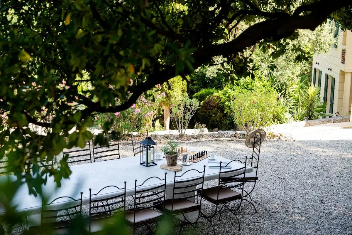 Outdoor dining area with a long white table, black chairs, a lantern, a potted plant, and a chess set, surrounded by trees and greenery.