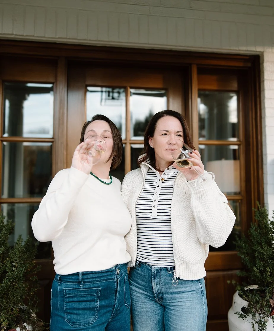 Two women are standing outside a house, each holding a glass of wine and drinking. They are dressed casually, with one wearing a white sweater and the other a striped shirt and white cardigan. They appear to be enjoying themselves.