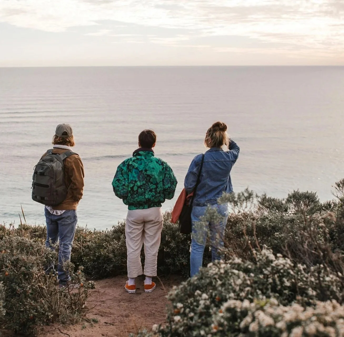 Three people standing on a dirt path by shrubs, looking out over the water at sunset or sunrise.