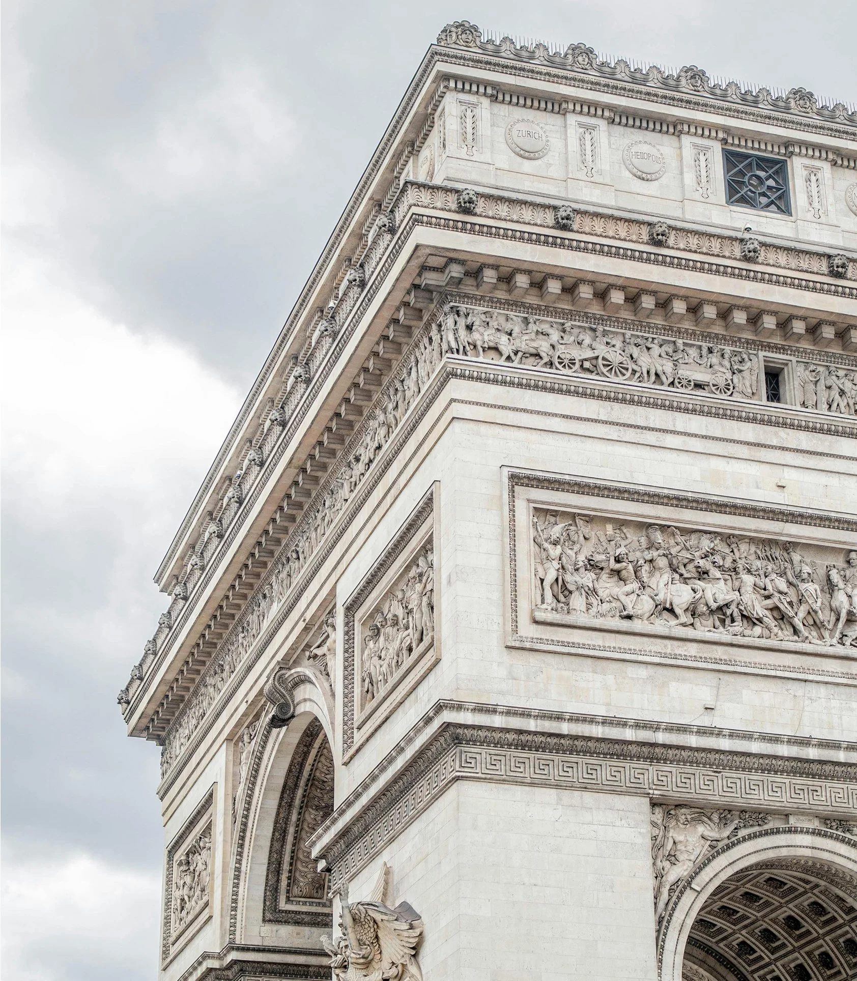 Close-up of the arch and upper part of the Arc de Triomphe in Paris, France, showcasing detailed sculptures, reliefs, and architectural elements with cloudy sky in the background.