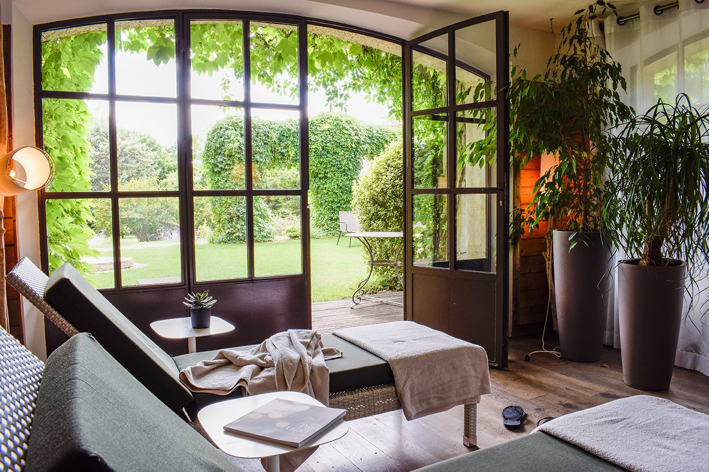 Living room with large open window doors leading to a green garden with trees and bushes. Inside, there are lounge chairs, a small table with a potted plant, a magazine, and large potted plants near the window.