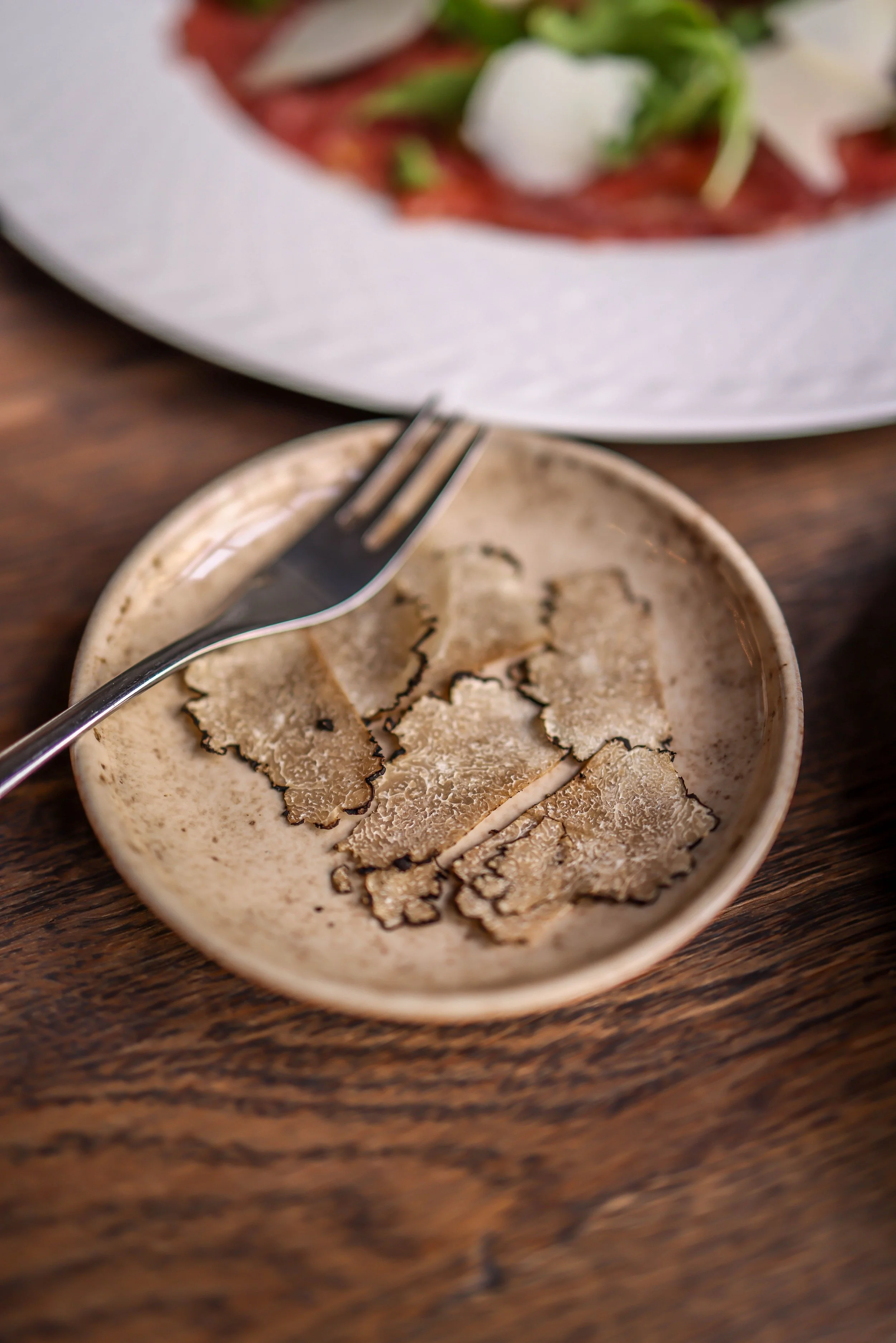Close-up of a small bowl with thin slices of truffle on a wooden table, with a fork resting on the bowl, and a blurred plate of food in the background.