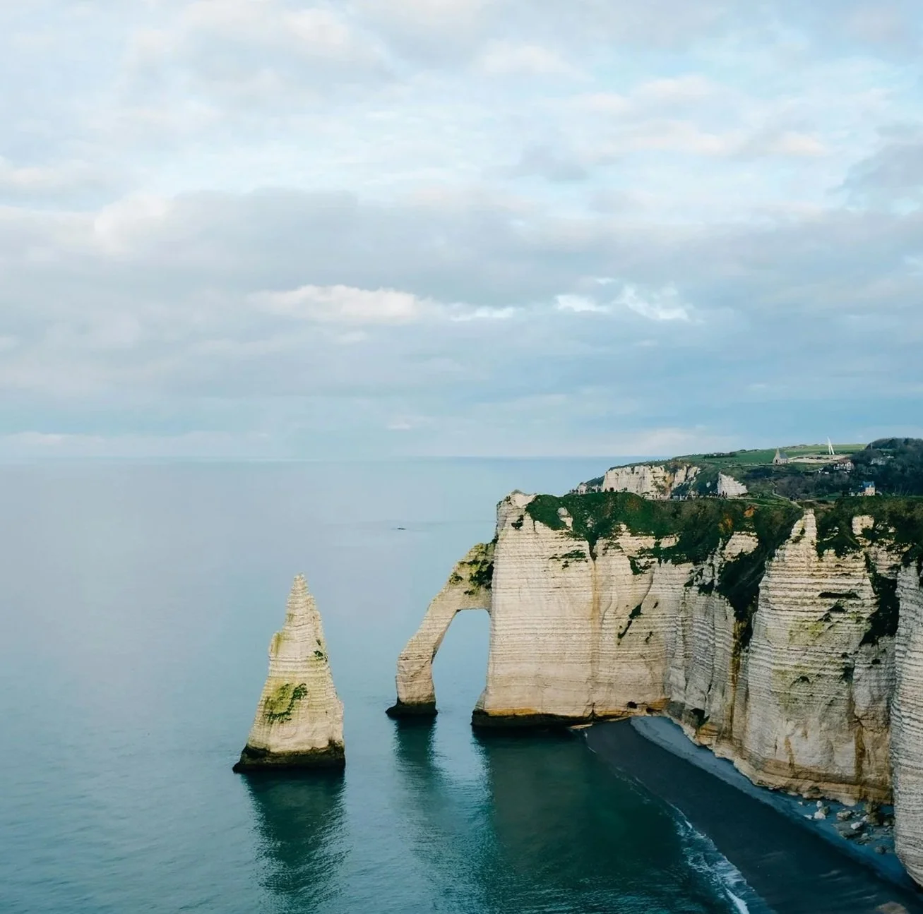 Cliffs along the coastline with a natural arch formation, overlooking the ocean, under a cloudy sky.
