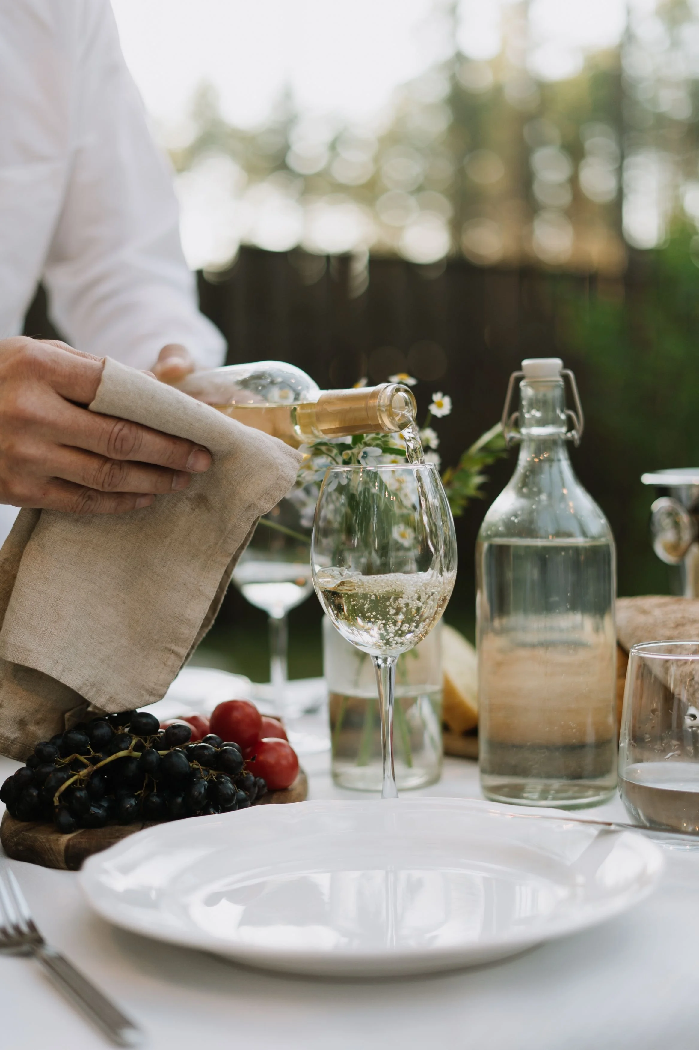 A person pouring white wine into a glass at an outdoor table with a blurred background of trees, with grapes and cherry tomatoes on a wooden board, and carafes of water on the table.