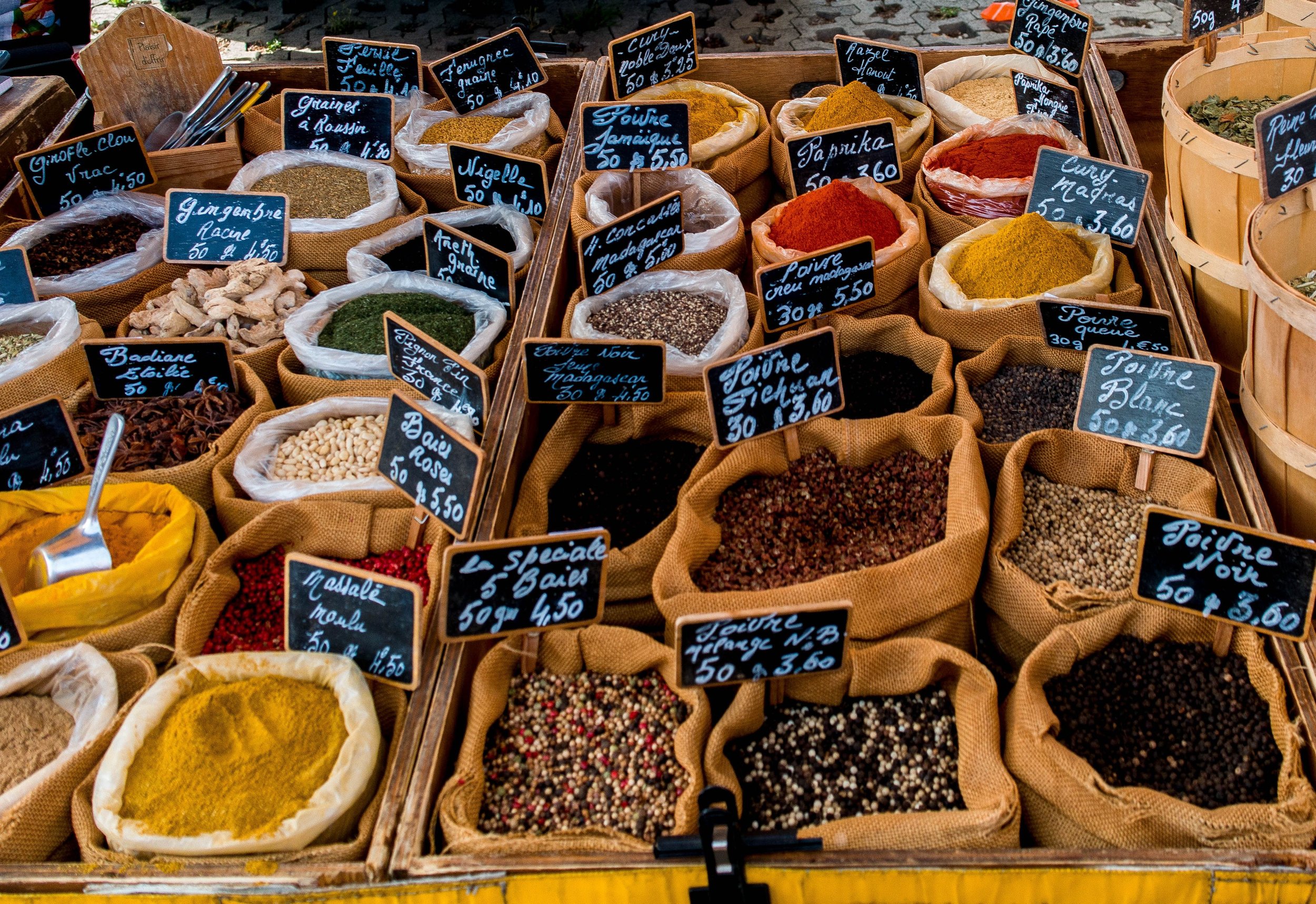 Assorted spices and herbs in burlap sacks at an outdoor market, each with handwritten black labels indicating the spice names and prices.