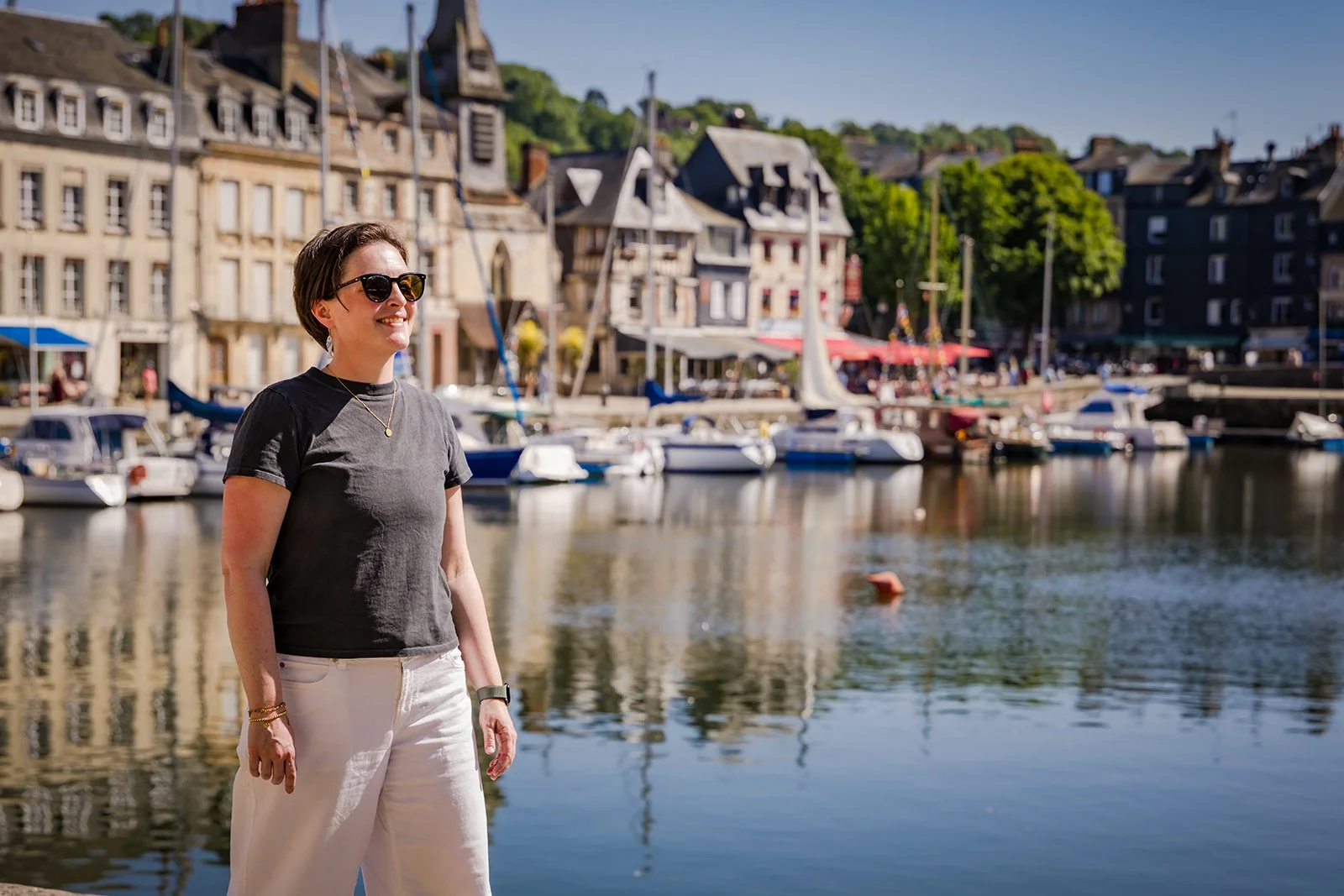 A woman standing by a marina with sailboats and yachts, smiling and wearing sunglasses, a gray T-shirt, and white pants.