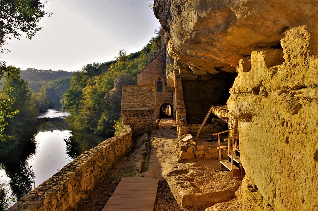 Scene of old stone building with a walkway along a river, surrounded by lush green trees during sunset.