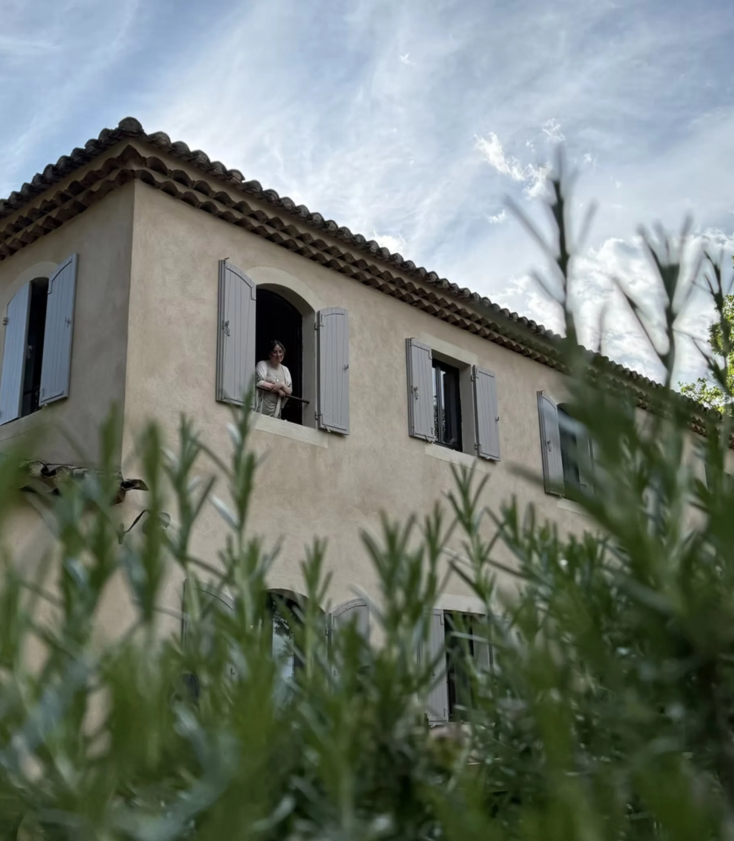 A woman standing by an open window of a beige house with gray shutters, with a cloudy sky above and green plants in the foreground.