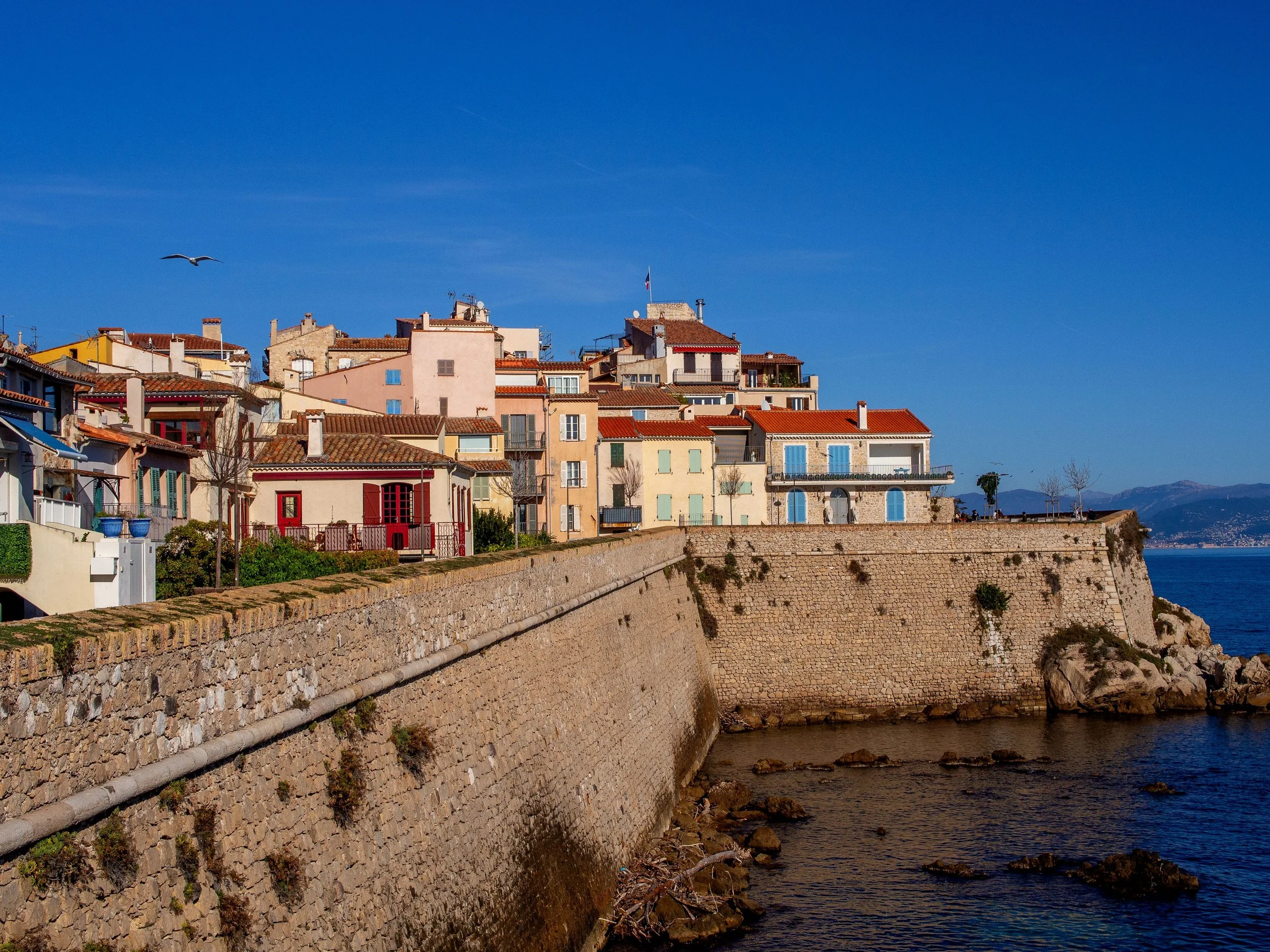 A coastal scene with a promenade and a stone seawall along the water, with colorful buildings with balconies and shutters, a bird flying overhead, and mountains in the background