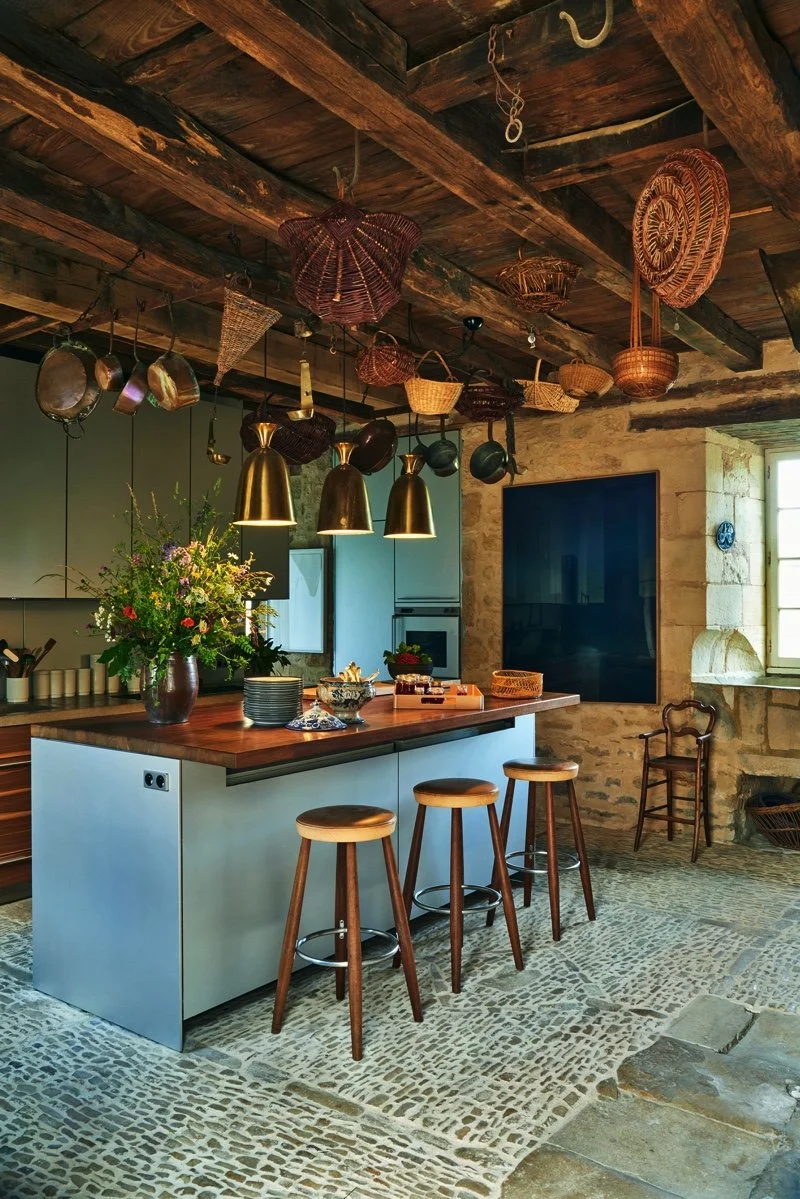 Rustic kitchen with wooden ceiling beams, hanging pots and wicker baskets, modern light fixtures over a kitchen island, and a stone wall with a window.