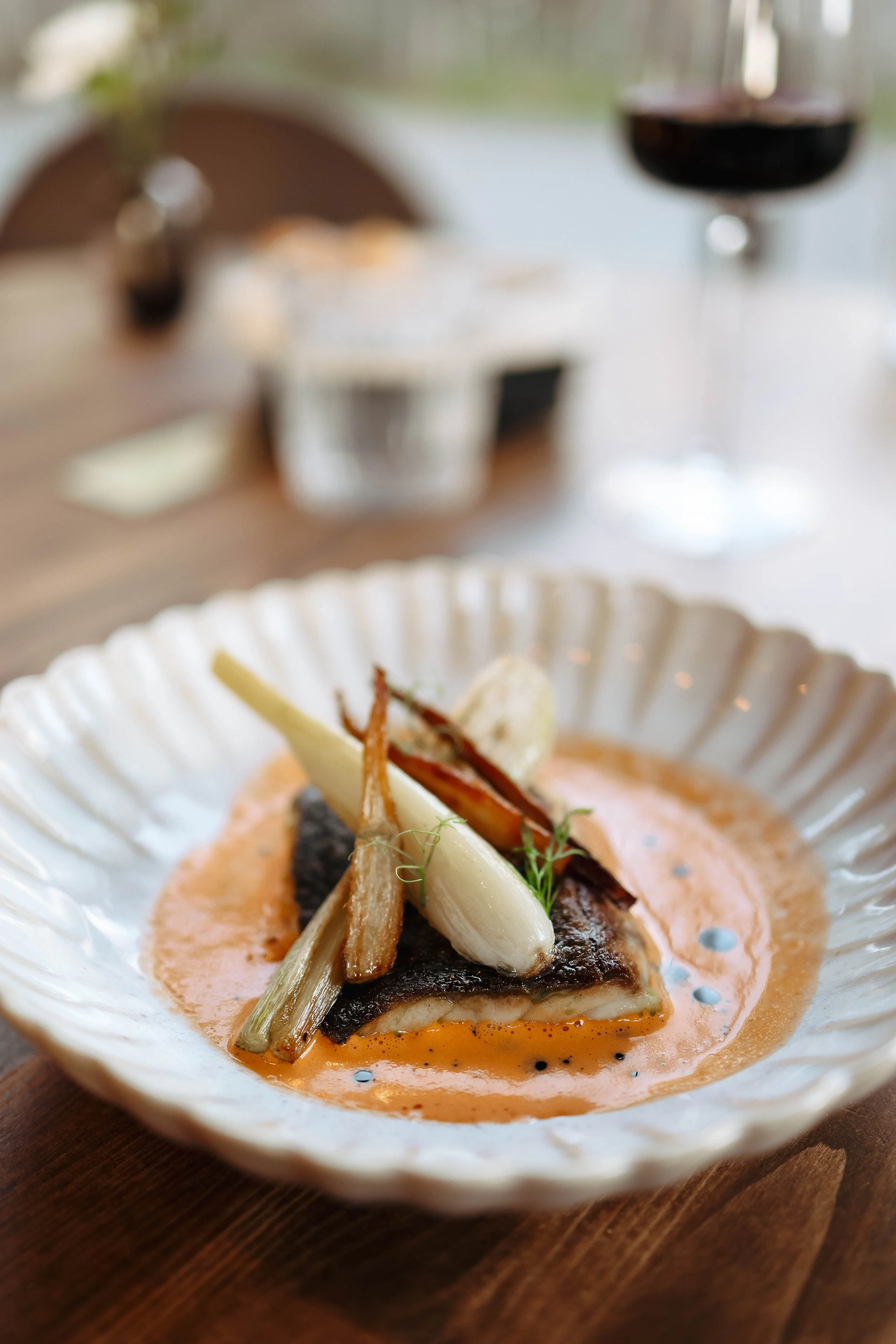 A plated dish featuring a seared piece of fish topped with grilled vegetables and herbs, served with a creamy orange sauce on a decorative white plate.