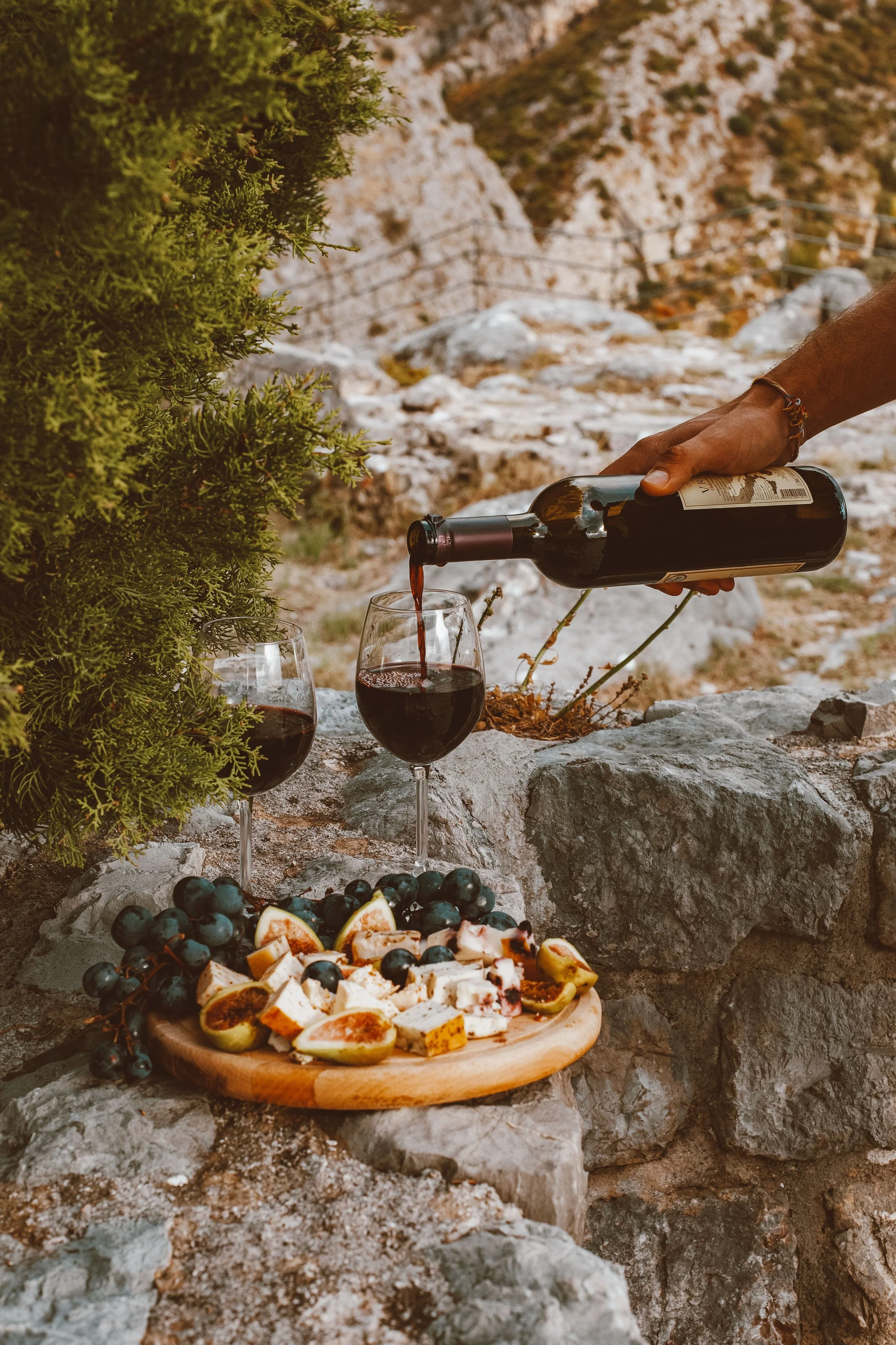 A hand pouring red wine into a glass, with a similar glass already filled, on a rocky surface outdoors. A platter of sliced figs, grapes, and cheese is in the foreground. There are green shrubs and rocky mountains in the background.