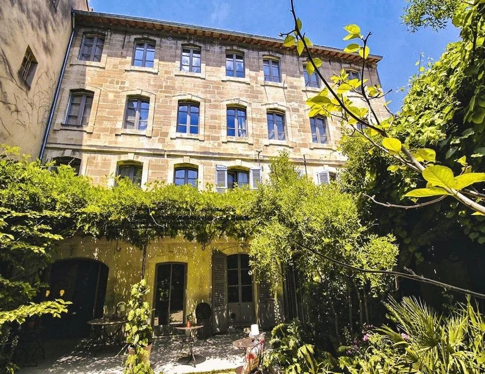 A stone apartment building with several balconies, surrounded by lush green trees and plants in a courtyard with outdoor furniture, under a bright blue sky.