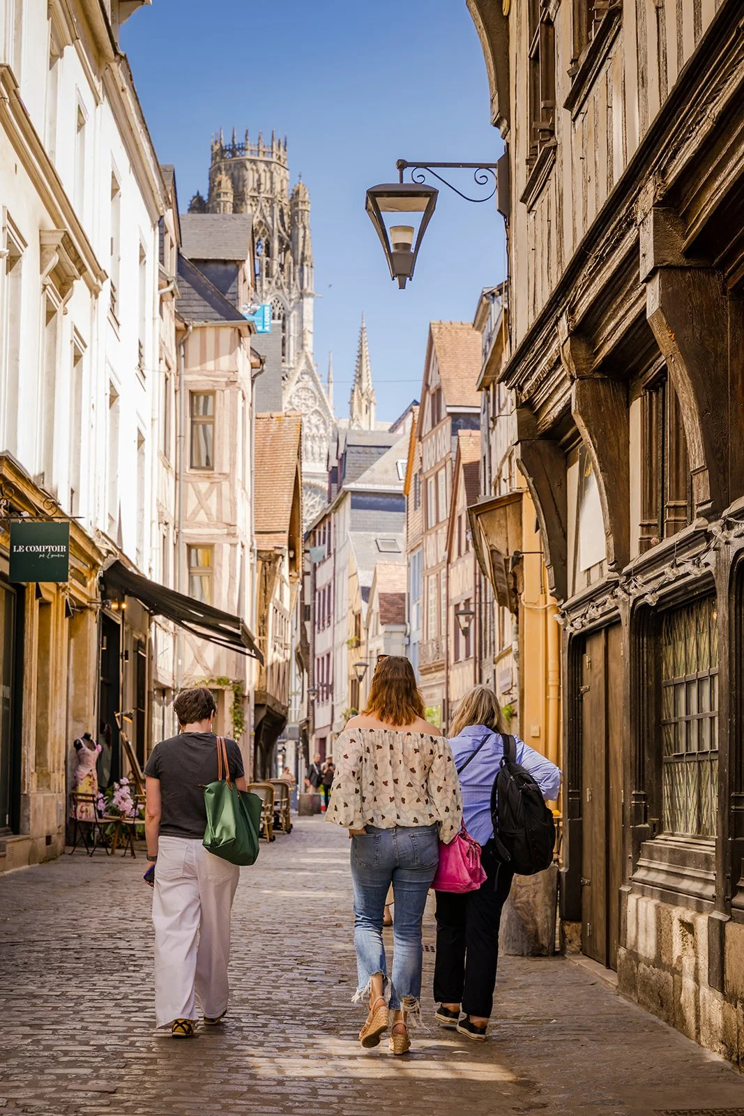 People walking down a cobblestone street in France with historic buildings, a Gothic cathedral in the background, and a clear blue sky.