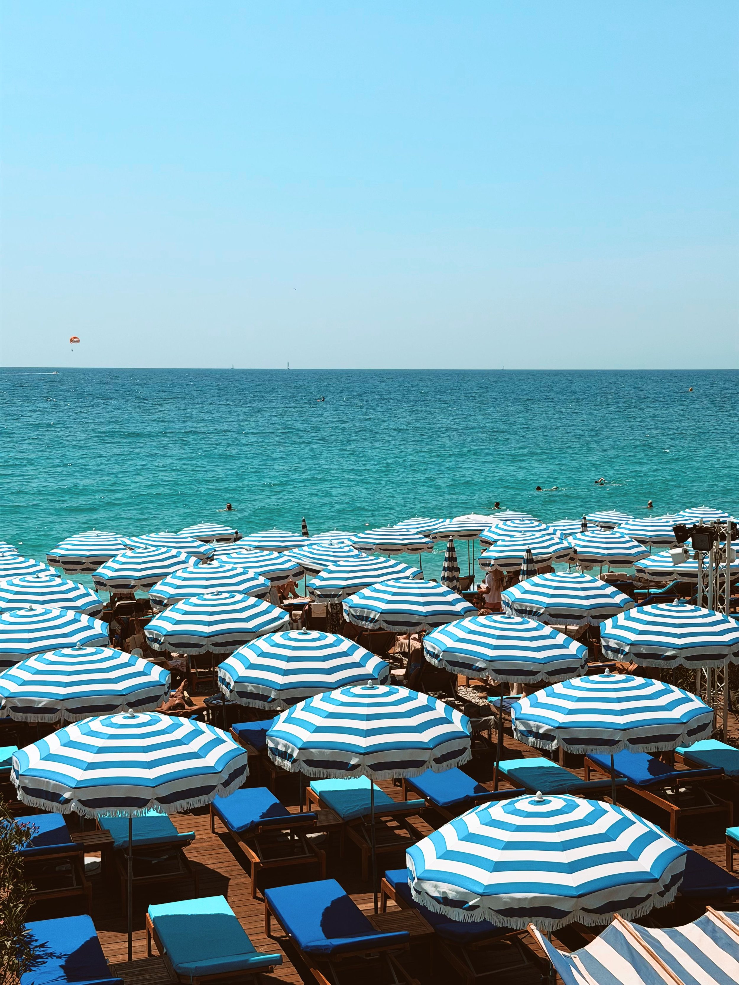 Beach scene with many blue and white striped umbrellas, people swimming and relaxing in the water, sailboats and parasailer in the distance under a clear blue sky.