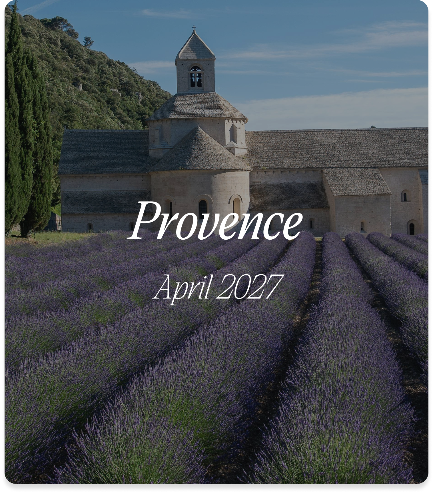 A stone church with a bell tower and cross on top surrounded by purple lavender fields, green trees, and a blue sky.