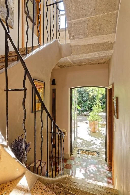 Open front door leading outside to a sunny garden area with trees and plants, view from an interior entryway with a tiled floor, framed wall art, and a decorative black metal railing staircase.
