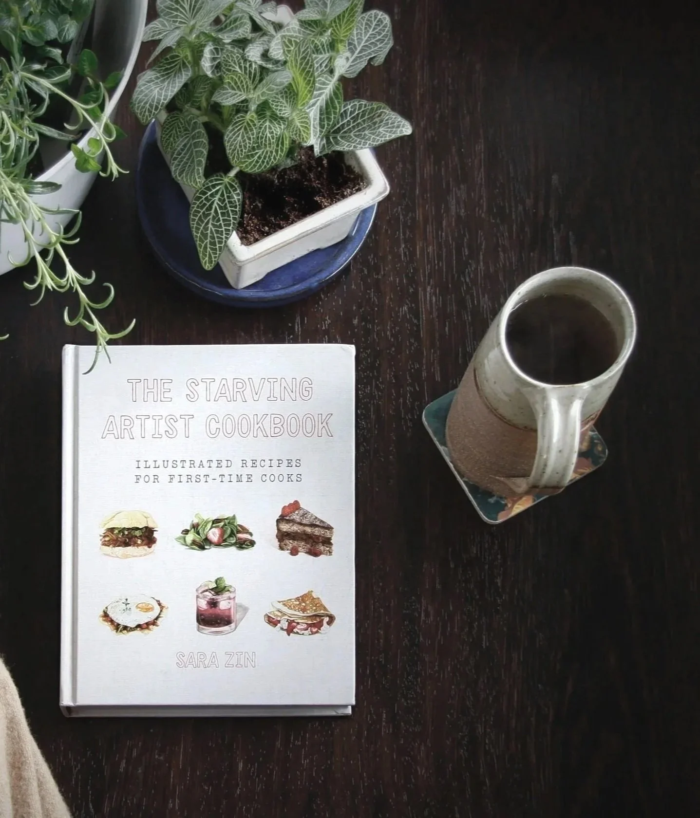 The cookbook titled "The Starving Artist Cookbook" by Sara Zin, with a mug of coffee on a dark wooden table.