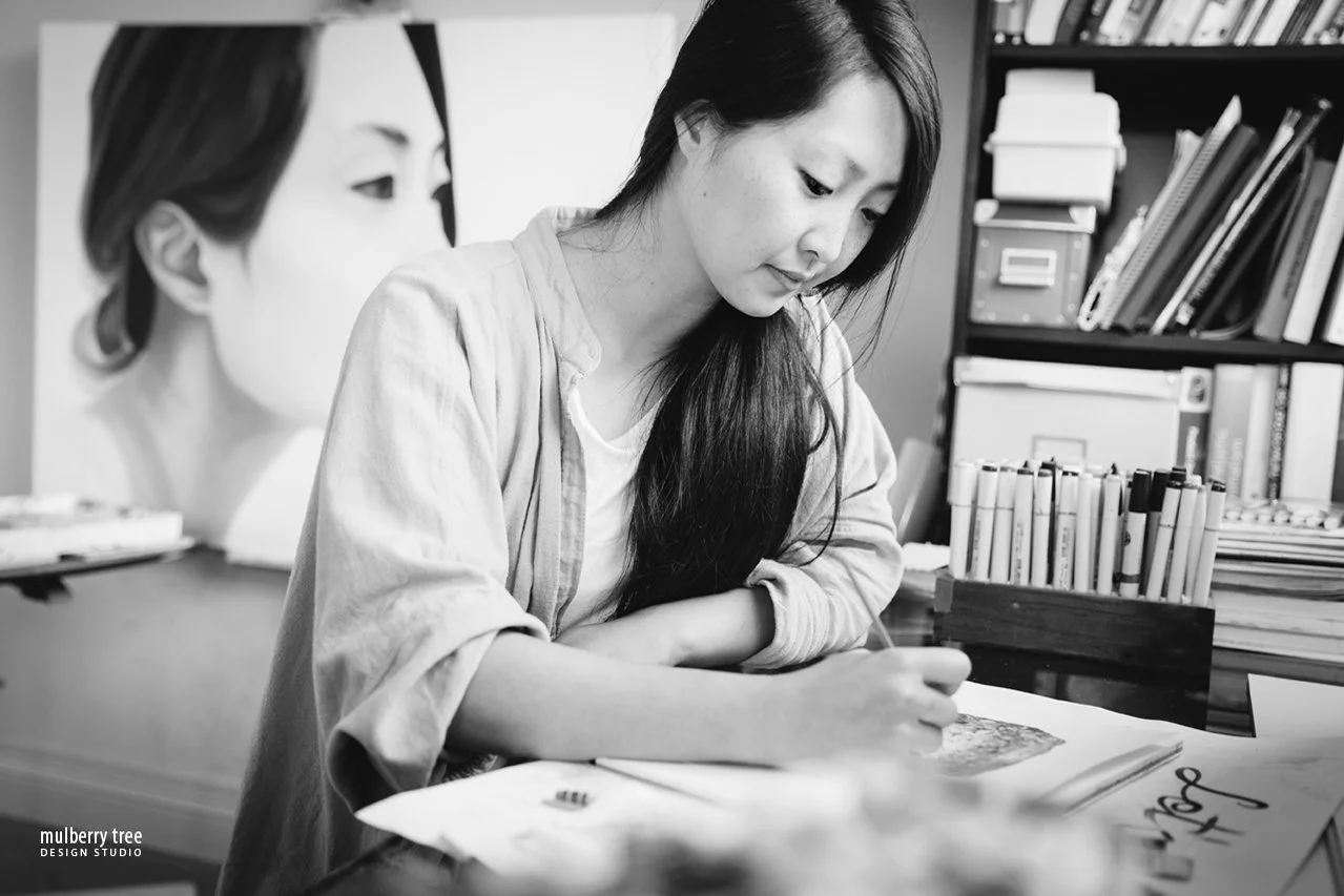 A photo of Sara Zin painting at a desk in a room with shelves filled with books and folders, and a large portrait painting in the background.
