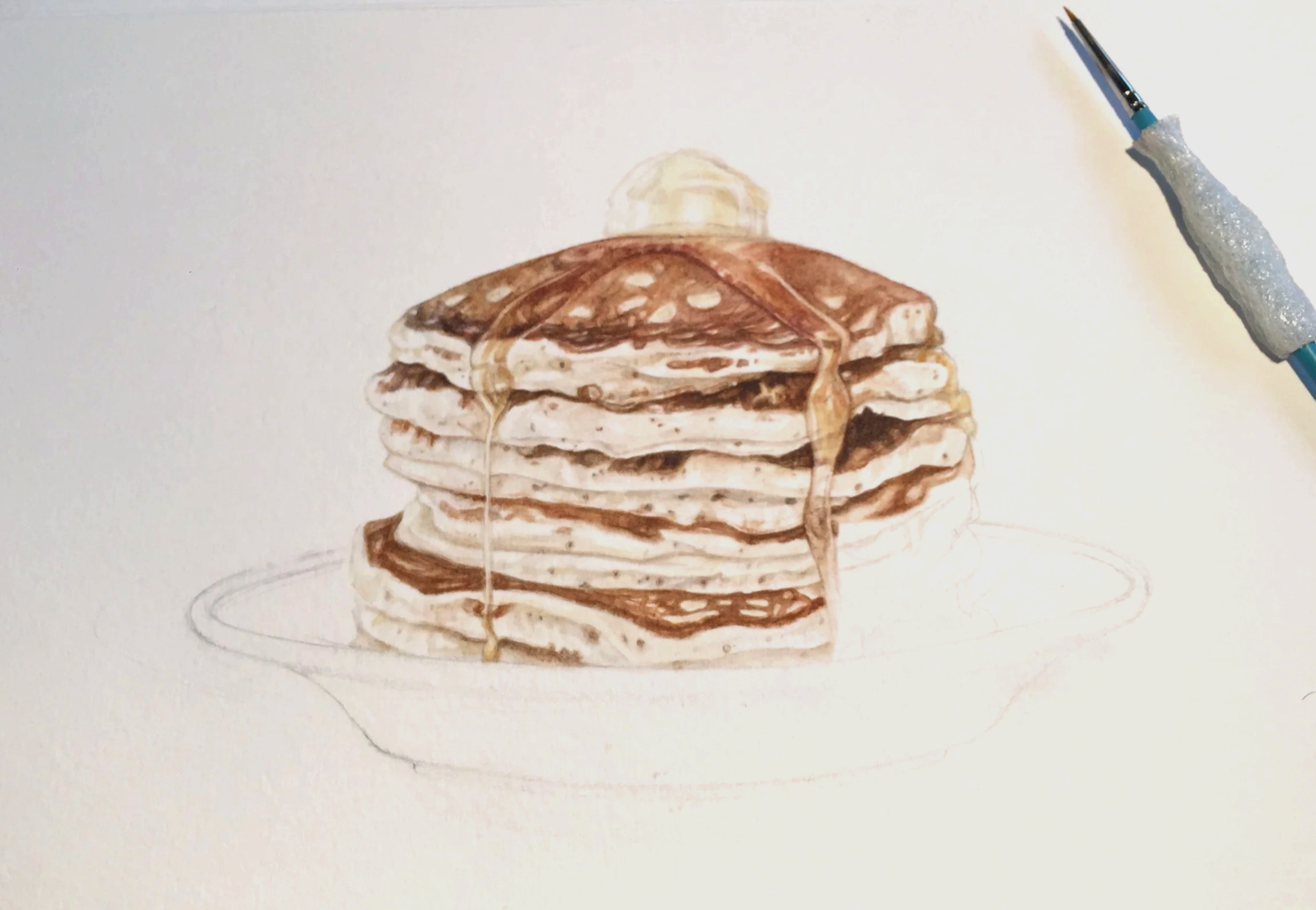 Work in progress photo of creating "The Starving Artist Cookbook" by Sara Zin. A watercolor painting of a stack of pancakes with syrup and a pat of butter on top, on a white surface with a watercolor paintbrush to the right.