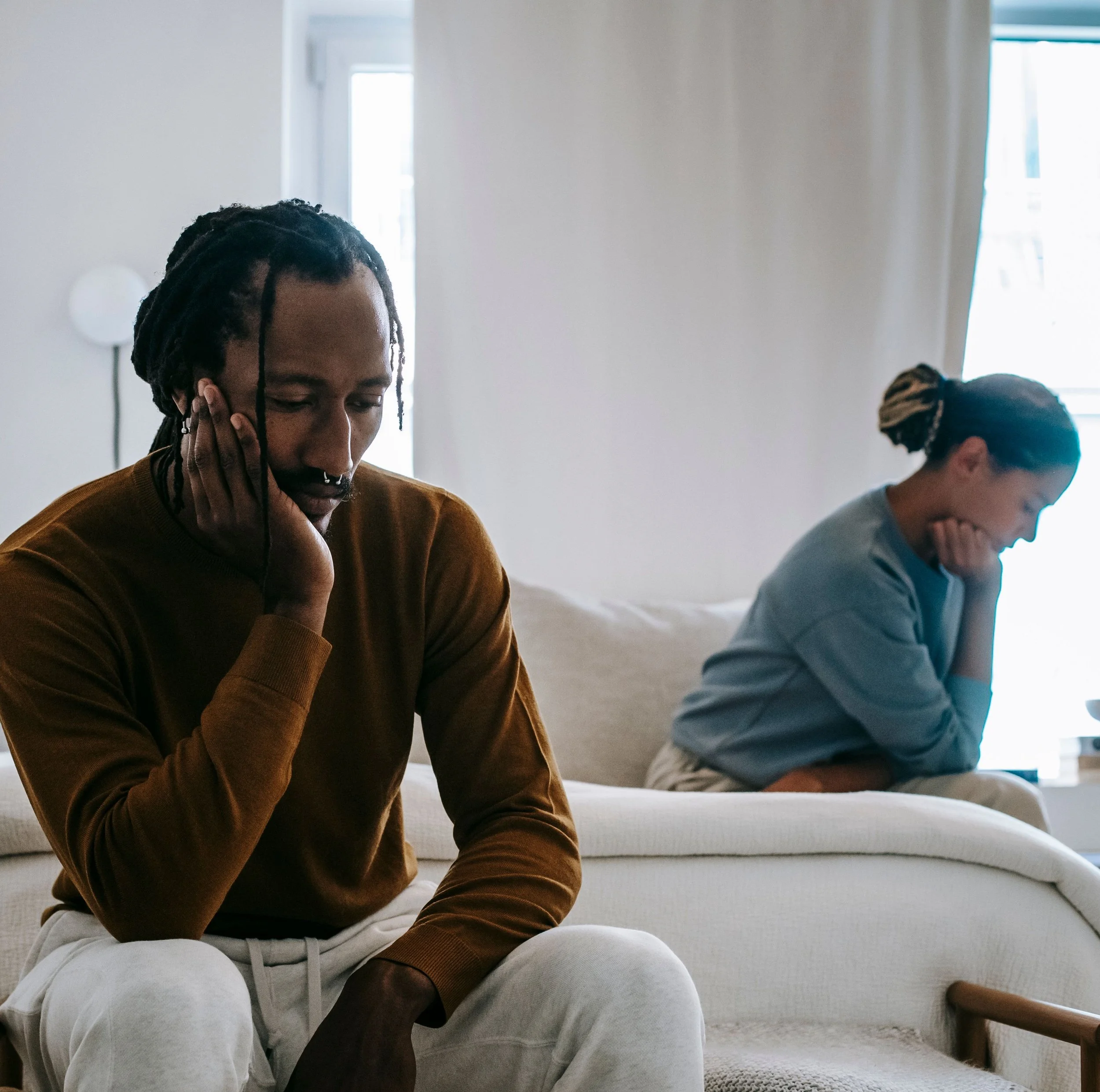 Two people, a couple,  sit on a beige couch in a bright room with large windows, one with dreadlocks and another with a head wrapped in a scarf, both appear to be resting or deep in thought.