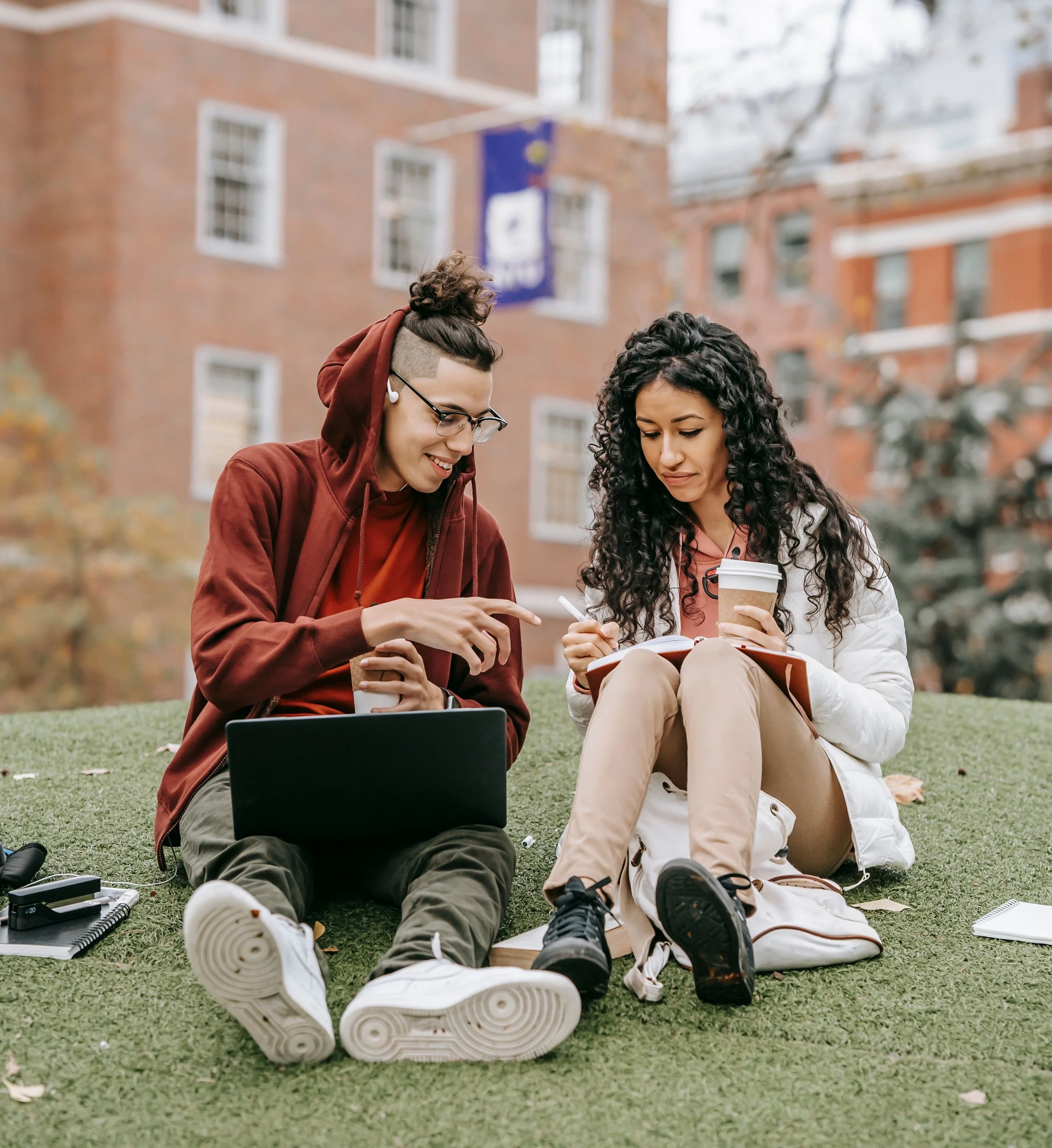 Two young women, college students, sitting on grass in front of a brick building, looking at papers and a notebook, with a laptop, coffee cups, and school supplies nearby.