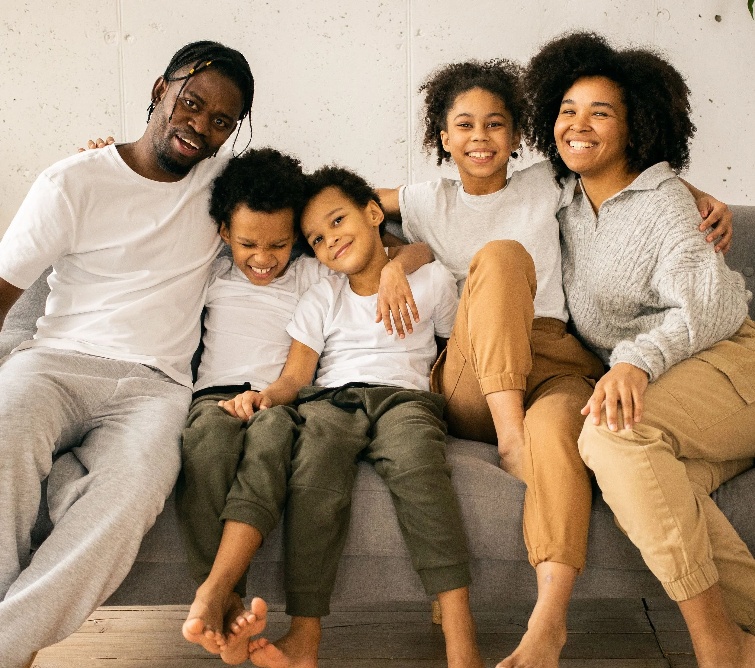 A diverse family of six sitting on a gray couch, smiling and enjoying each other's company. The family includes a man, woman, and four children with various hairstyles and expressions.