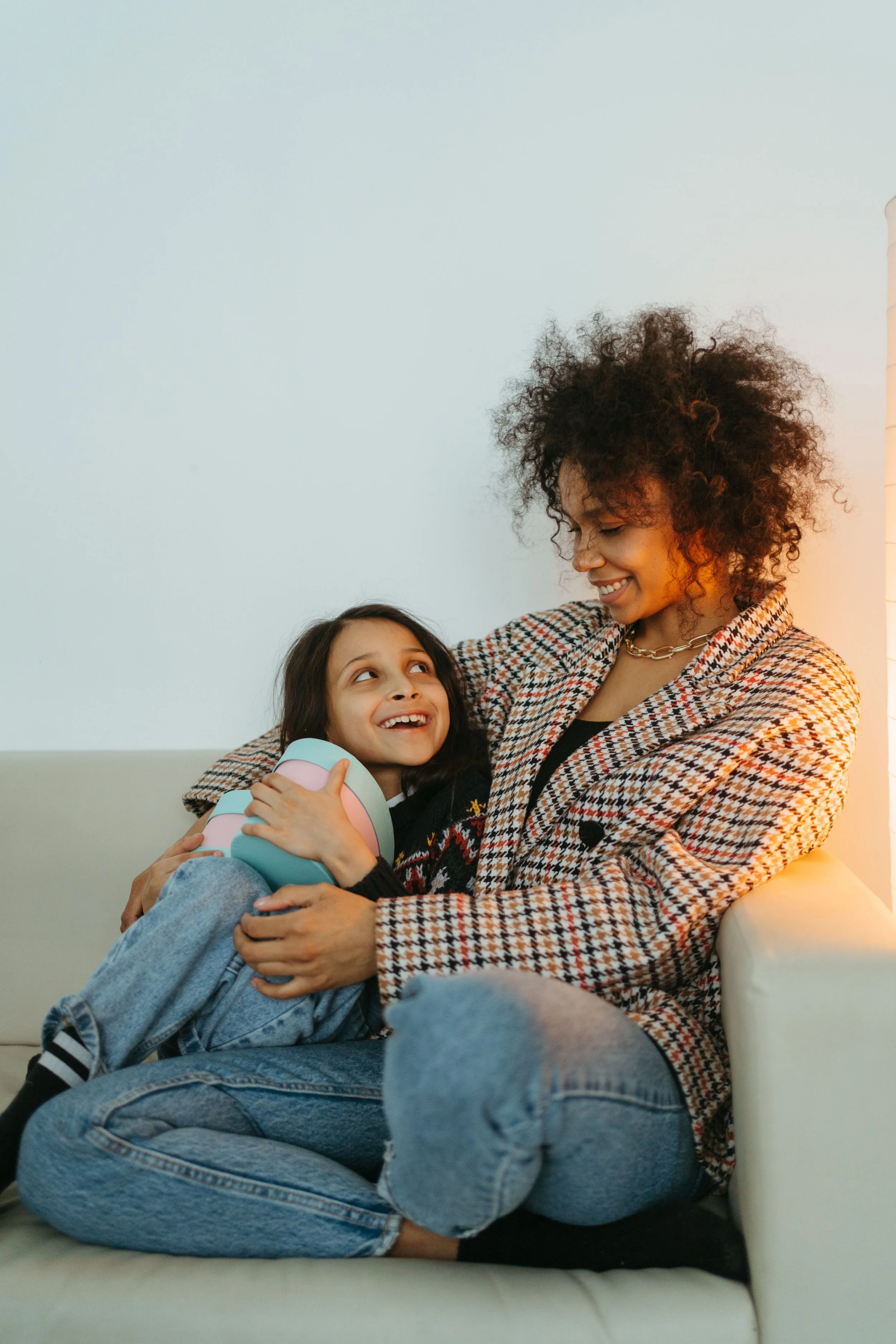 A woman, the mother, with curly hair and a smiling face wearing a plaid coat sitting on a white couch, holding a young girl, the daughter,  with long dark hair, who is looking up at her and holding a toy. The setting is indoors with a white wall.
