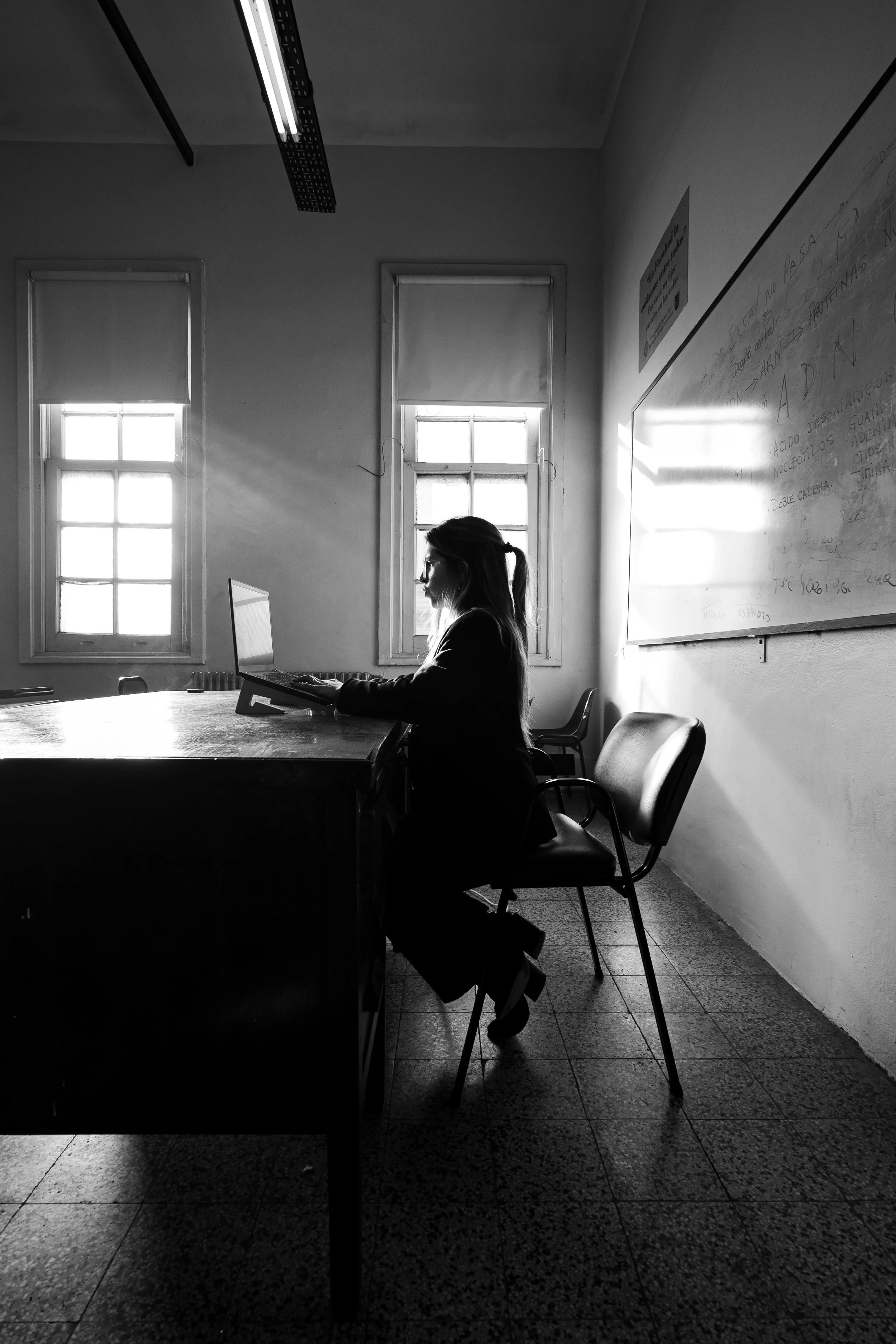 Black and white photo of a female teacher sitting at a desk with a laptop, illuminated by natural light coming through two windows behind her, with a whiteboard mounted on the wall to the right.