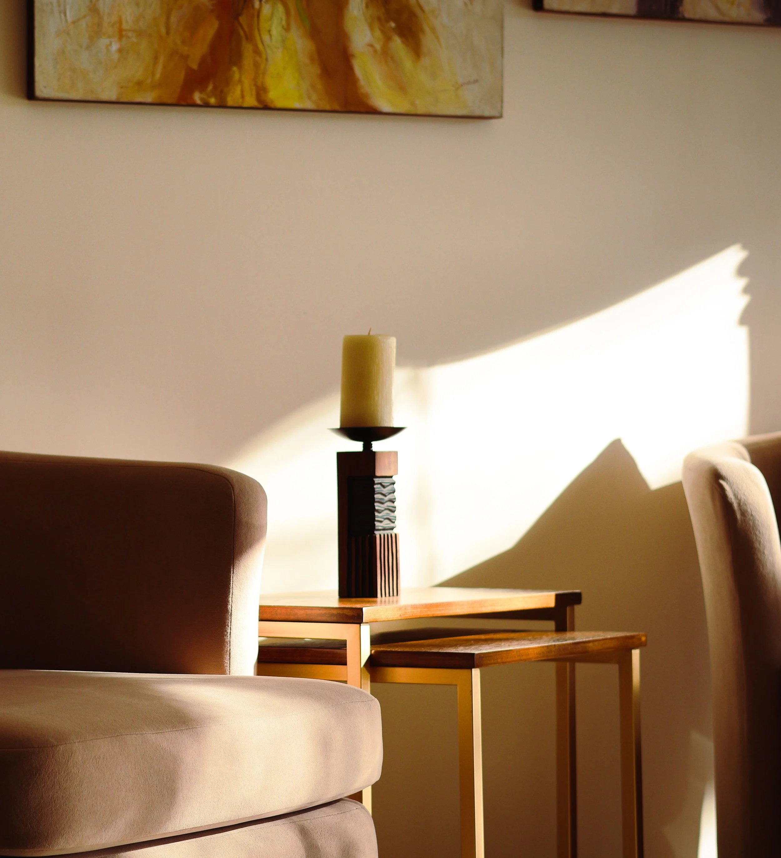A beige cushioned chair in the foreground on the left side, a wooden table with a decorative candle holder in the center, a wooden staircase with multiple steps in the background, a painting with warm colors on the wall, and sunlight casting shadows on the walls.