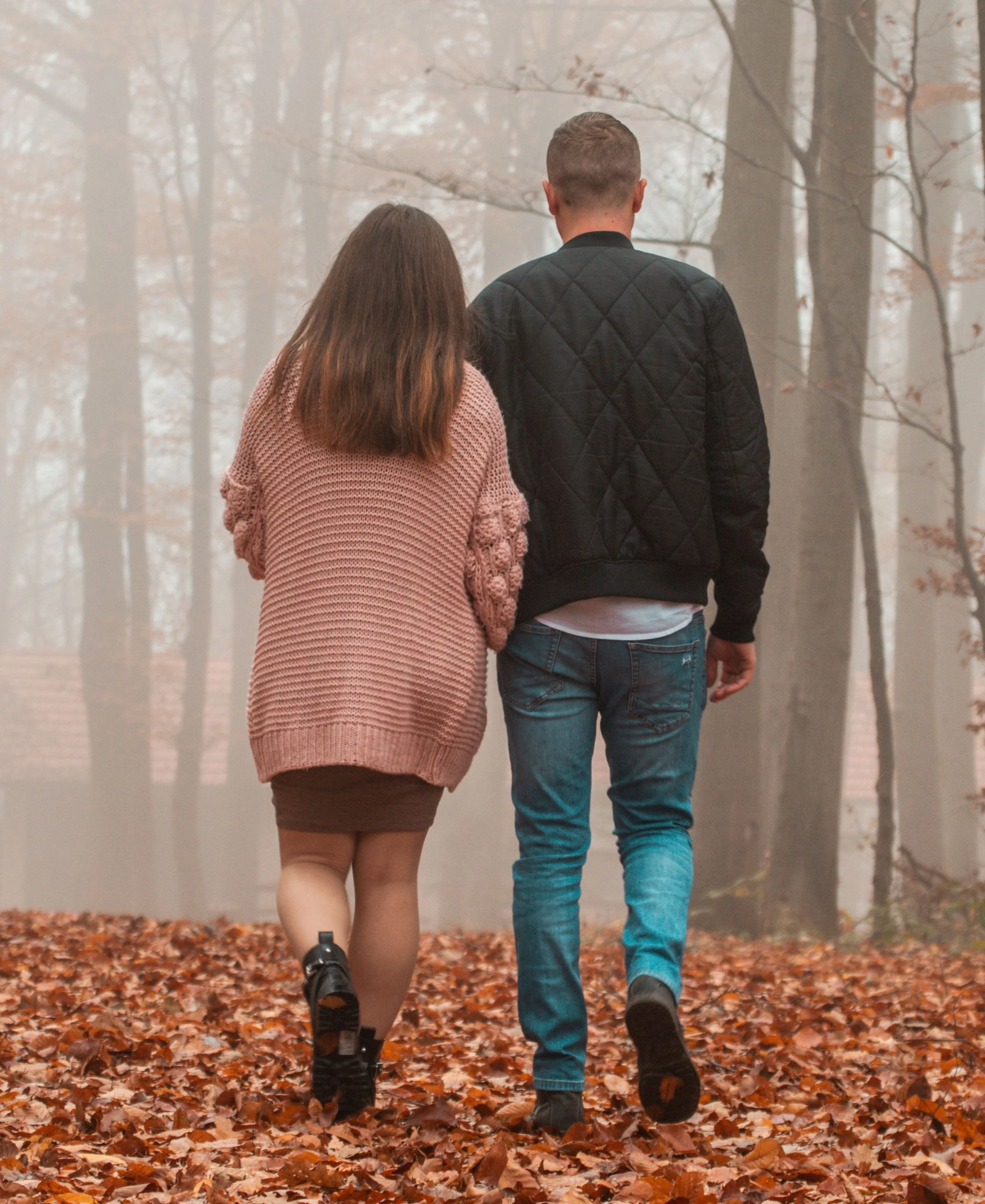 A couple walking together in a forest during autumn, surrounded by fallen leaves and trees with sparse branches.