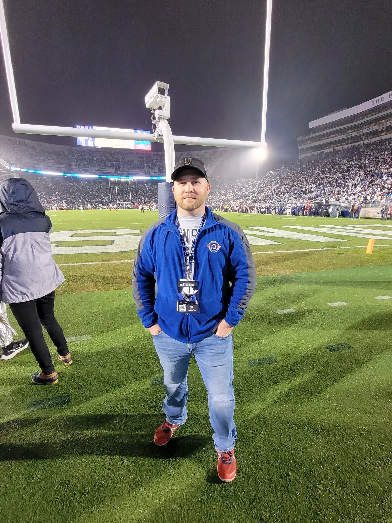 A man standing on a football field at night, wearing a black cap, blue jacket, jeans, and red sneakers, with hands in his pockets, in front of empty goalposts with stadium filled with spectators in the background.