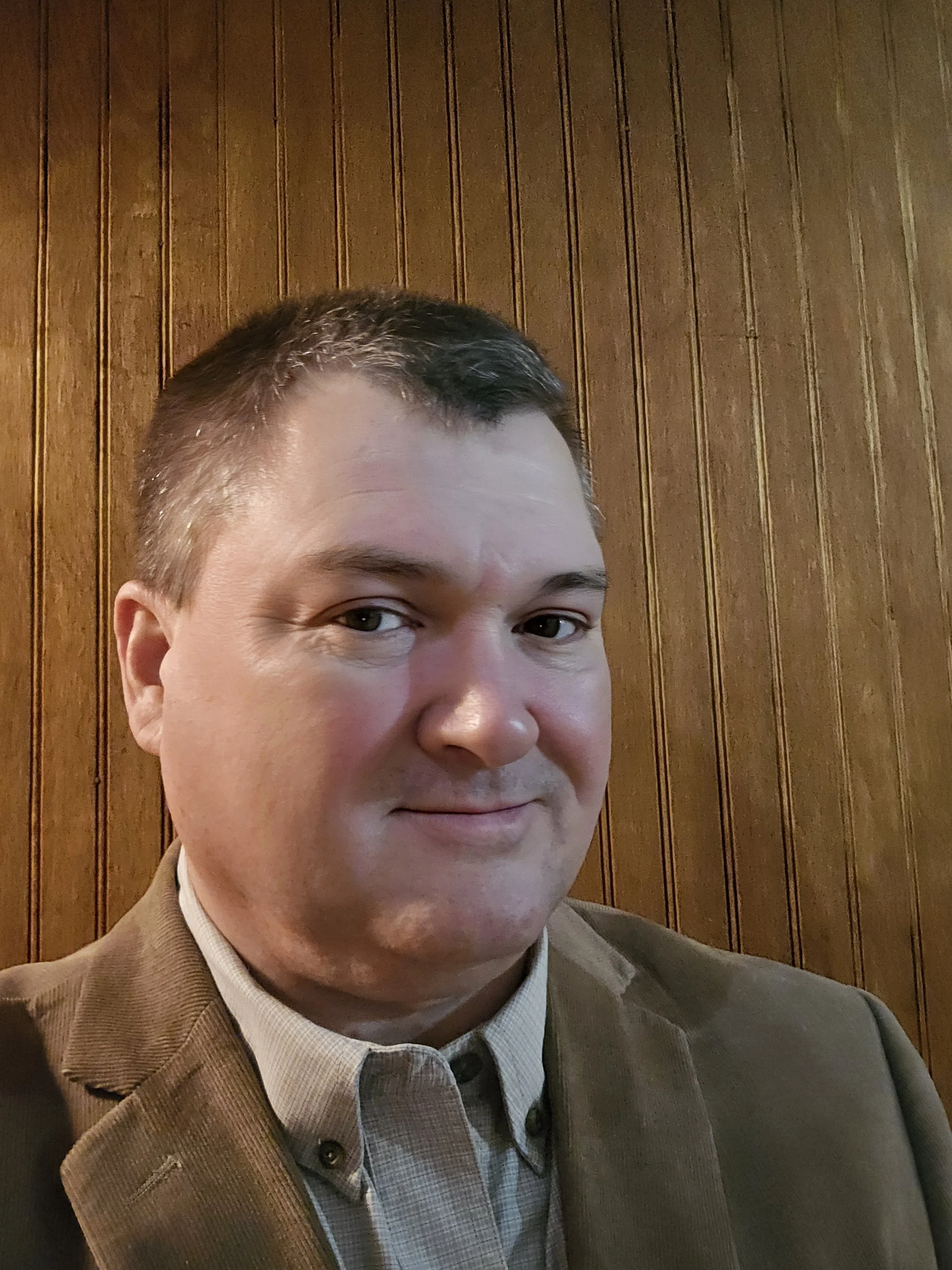 Close-up of a man with short brown hair, light complexion, wearing a brown blazer and a light-colored button-up shirt, sitting in front of a wooden paneled wall.