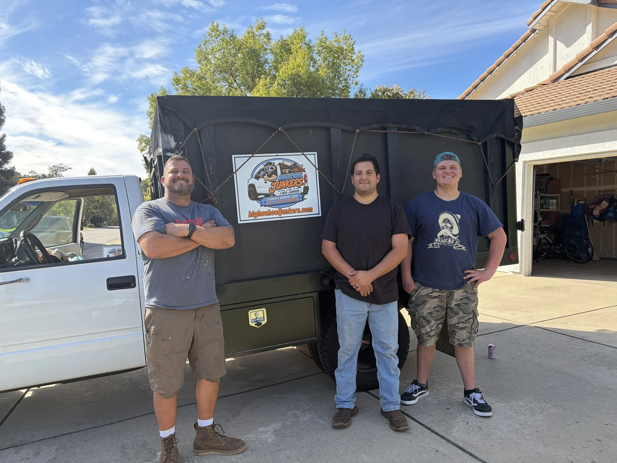 Three men standing in front of a black truck with High School Junkers logo, outside a house with a garage, on a sunny day.